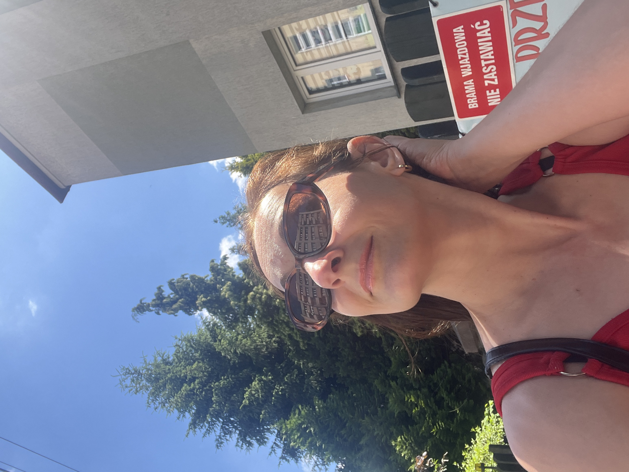 In a medium close-up outdoor shot, a woman with auburn hair, wearing sunglasses and a red top, looks up towards the sky. Her hand is raised to her head, and she is touching her earlobe. The background features a building, trees, and a sign that reads "BRAMA WJAZDOWA NIE ZASTAWIAĆ". The sky is blue with some clouds. The reflection in the woman's sunglasses shows a multi-story building.