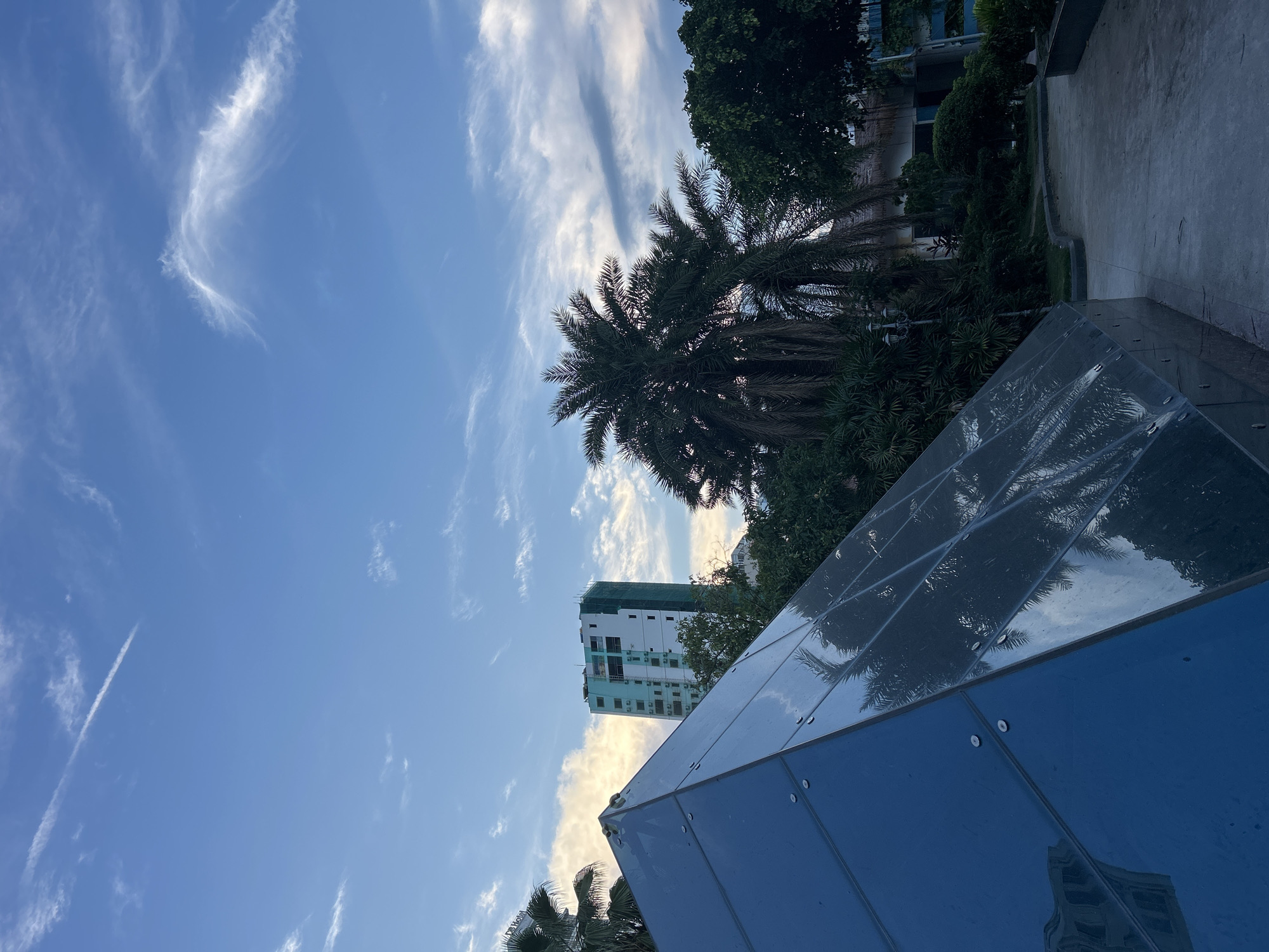 The image captures a view of a city park area in Nha Trang Ward, Vietnam, featuring a modern glass pyramid structure in the foreground, reflecting palm trees and the sky. Behind the pyramid, lush green foliage and palm trees are visible, with a tall, light-colored building under construction in the mid-ground, draped in green netting. The sky above is a clear blue, dotted with wispy white clouds, including a prominent contrail from an aircraft. A paved pathway curves through the park, lined with trees and some low-lying bushes. The lighting suggests it is either early morning or late afternoon, with a soft, diffused light. No people are present in the scene.