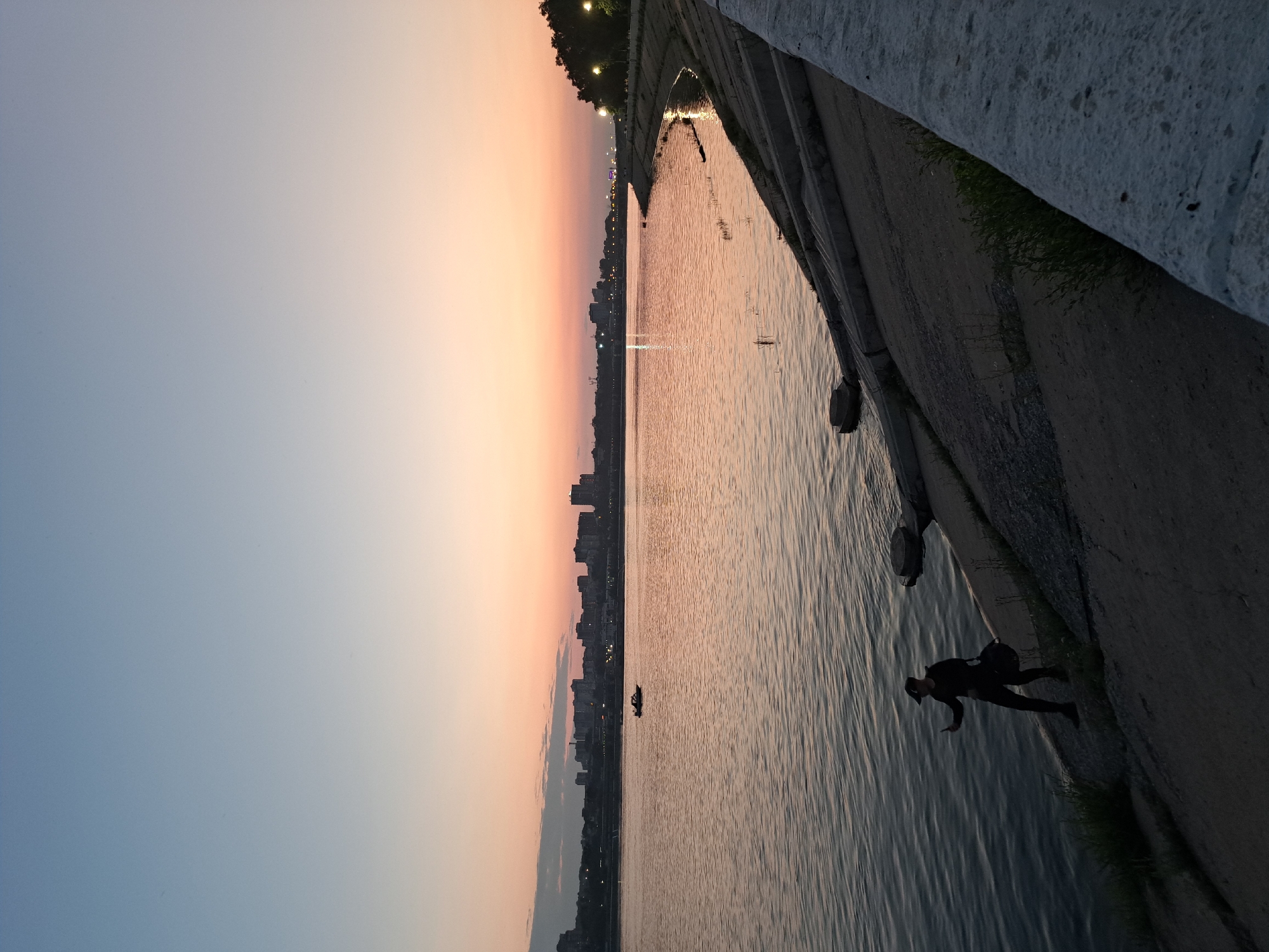 The image depicts a sunset scene over the Angara River in Irkutsk, Russia. In the foreground, a person dressed in dark clothing, wearing a cap and carrying a dark bag, stands on the embankment, holding up a mobile phone, seemingly taking a photo or video. The embankment is made of concrete and stone, with patches of grass growing through. The river itself appears calm, with gentle ripples reflecting the soft colors of the twilight sky, which transitions from pale blue at the top to hues of orange and pink near the horizon. Across the river, the city skyline of Irkutsk is visible, characterized by a cluster of buildings and lights. A small boat is present on the water in the mid-ground. The overall atmosphere is tranquil, with the fading light creating a serene ambiance.