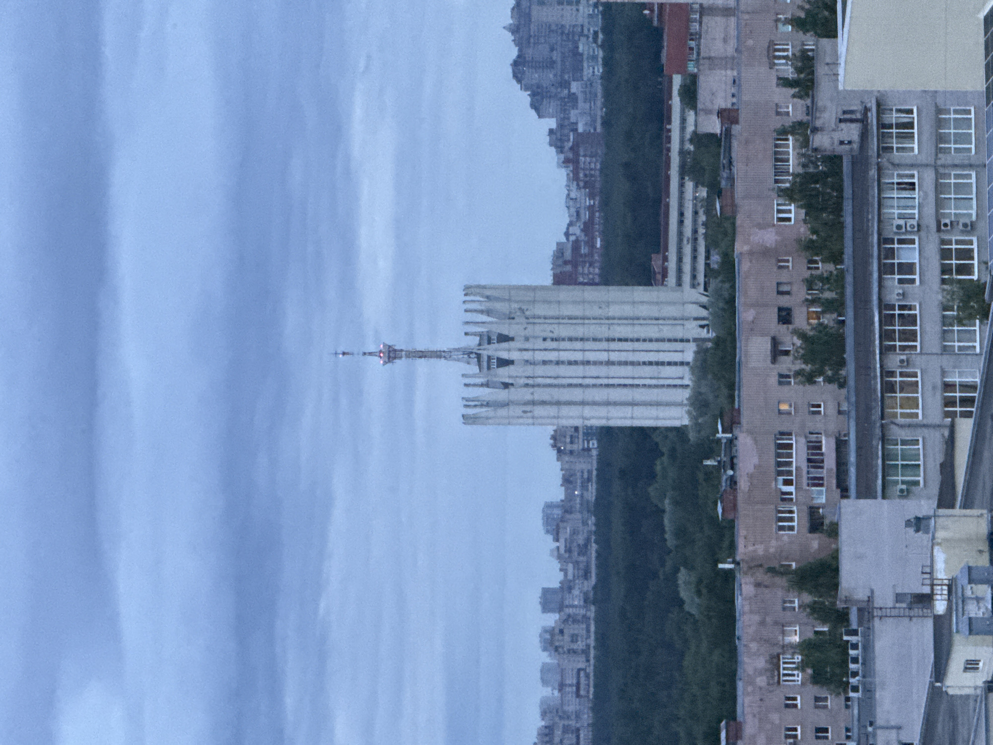 The image is a medium-distance aerial view of an urban landscape under a cloudy sky. In the foreground, there are several apartment buildings with visible windows and balconies. Behind these, a dense line of dark green trees forms a natural barrier. Beyond the trees, a striking white, vertical structure dominates the mid-ground. This structure has a distinctive design with pointed vertical elements along its top and a slender antenna or spire at its peak, which has red lights. Further in the background, a city skyline with numerous multi-story buildings is visible, stretching across the horizon. The overall lighting suggests either early morning or late evening, with muted colors and soft shadows. The image appears to be taken from a high vantage point, possibly another building or drone.