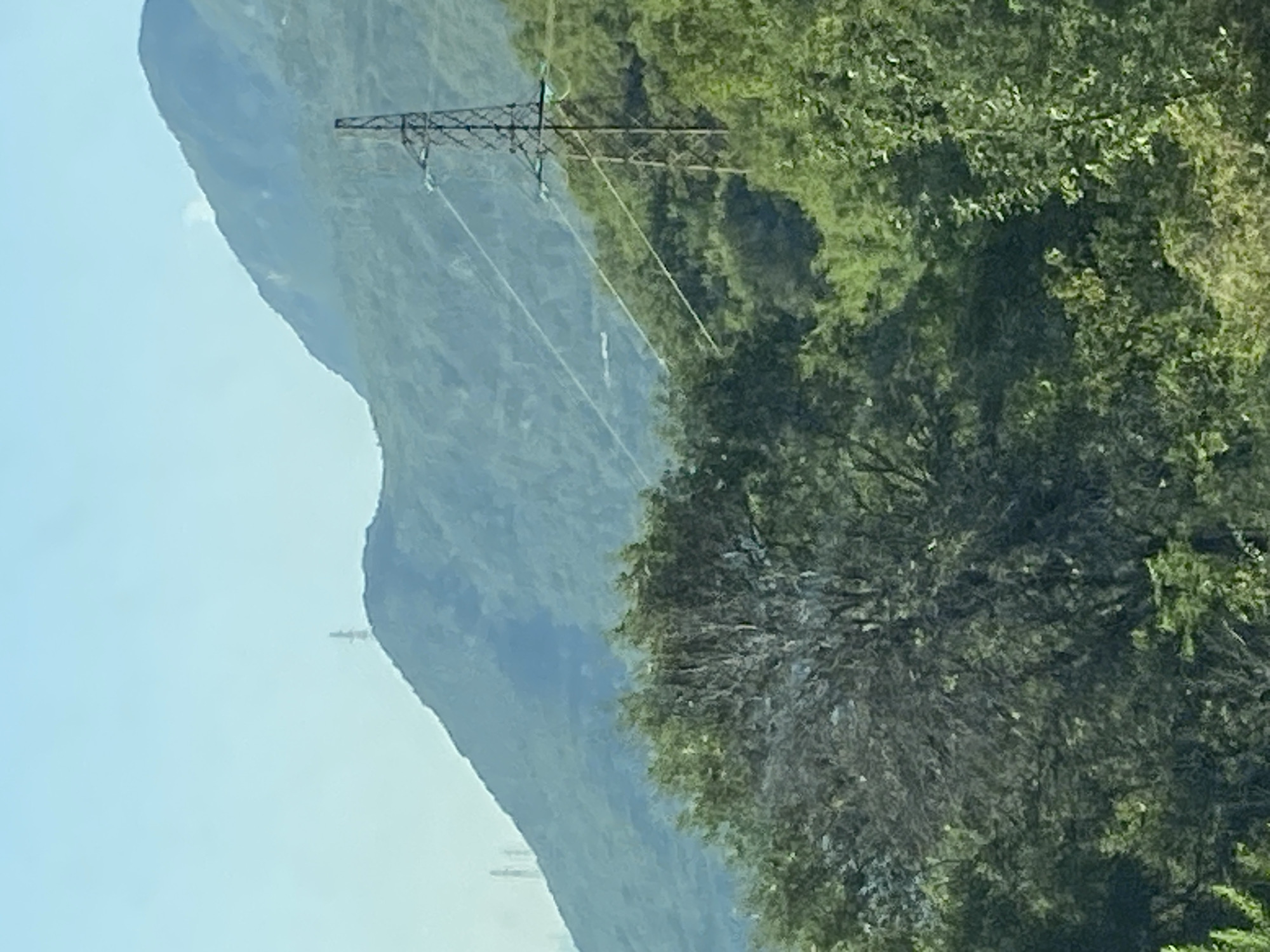 The image depicts a mountainous landscape with a power line tower in the foreground, situated in the city of Seoce, Montenegro. The tower is a tall, lattice structure supporting electrical wires that stretch across the frame. Dense green trees and foliage cover the lower portion of the image, obscuring the ground. In the background, rolling hills and mountains rise against a clear blue sky. Several other power line towers are visible in the distance, scattered across the slopes of the mountains. The scene appears to be a daytime shot, with natural lighting illuminating the landscape. There are no people or animals visible in the image.