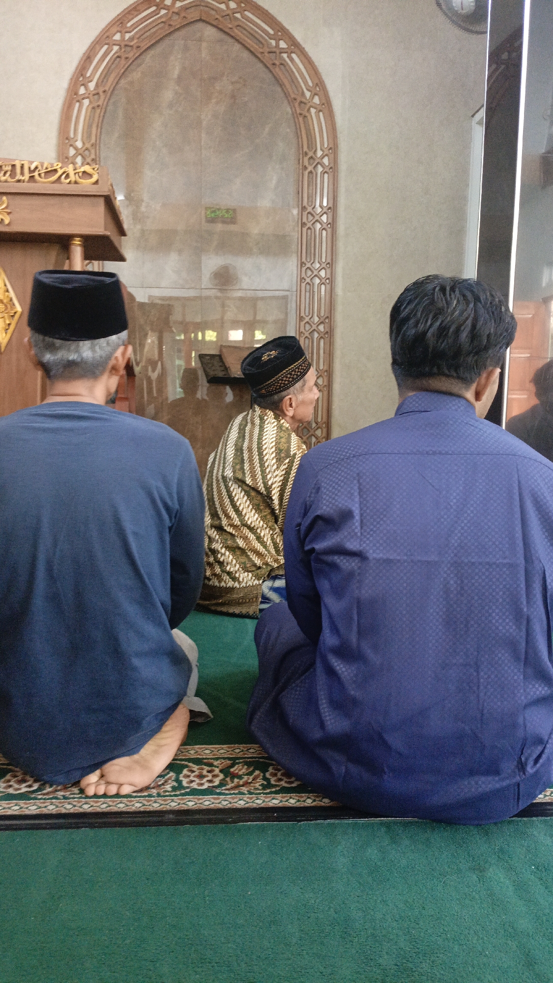 Three men are seated on a green carpet inside a building that appears to be a mosque. The man on the left, viewed from behind, wears a dark blue long-sleeved shirt and a black cap. His bare feet are visible. The man in the center, also viewed from behind, wears a patterned brown and white shirt and a black cap. The man on the right, viewed from behind, wears a dark blue patterned long-sleeved shirt and a black cap. The background features an arched doorway with intricate wooden latticework and a marble-patterned wall. A wooden podium is partially visible on the left. The overall impression is of a religious gathering or prayer session. The image context suggests this scene is located in Salatiga, Indonesia.