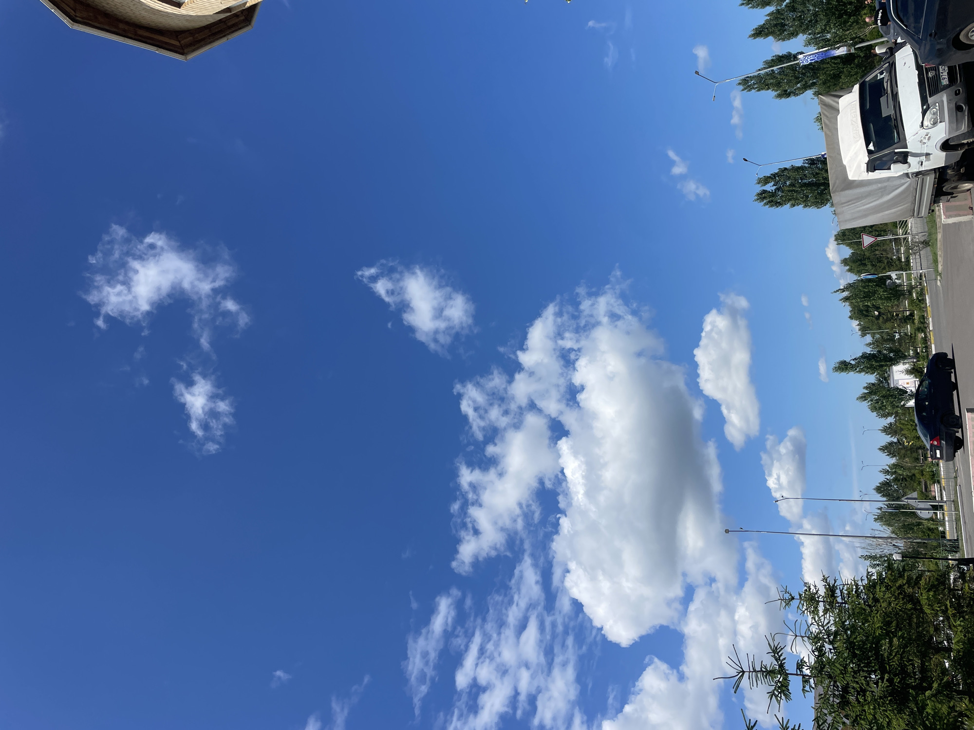 A bright blue sky with scattered white clouds dominates the upper portion of the image. In the far upper right corner, a portion of a brown, cylindrical building with a light-colored roof is visible. The lower part of the image depicts a street scene with several vehicles and trees. On the left, a dark green coniferous tree is prominent. A black sedan is parked on the right side of a road. Further to the right, a white truck with a gray tarp-covered cargo area is parked. Several tall, slender trees line the background, and lampposts are visible. The overall impression is a sunny day in an urban or semi-urban environment.