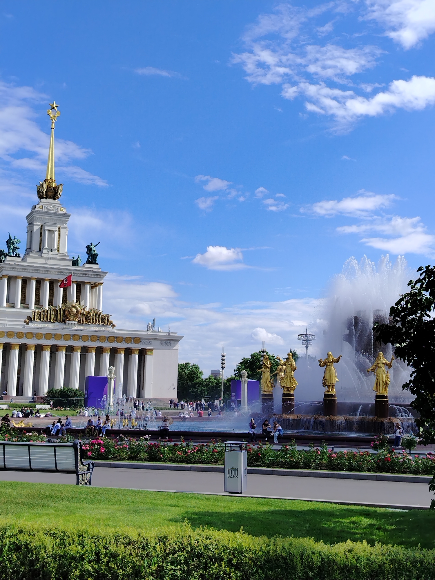 This image depicts a view of the "Friendship of Nations" fountain and the main pavilion at VDNKh (Exhibition of Achievements of National Economy) in Moscow, Russia, on a sunny day with scattered clouds. The fountain, a central feature, is adorned with gilded statues of women representing the former Soviet republics. Water cascades from multiple tiers, creating a dynamic spray. To the left, the imposing structure of the main pavilion, also known as Pavilion No. 1, stands prominently. Its facade is characterized by classical architecture, featuring a colonnade, ornate detailing, and a tall spire topped with a star. Several people are visible in the foreground and middle ground, some sitting on benches or near the fountain, while others are walking. The area is landscaped with green lawns and flowerbeds, predominantly featuring pink and red roses. A park bench is positioned in the lower left of the frame, and a trash receptacle with Cyrillic lettering is visible near the fountain. The overall scene captures a popular public space within the VDNKh complex.