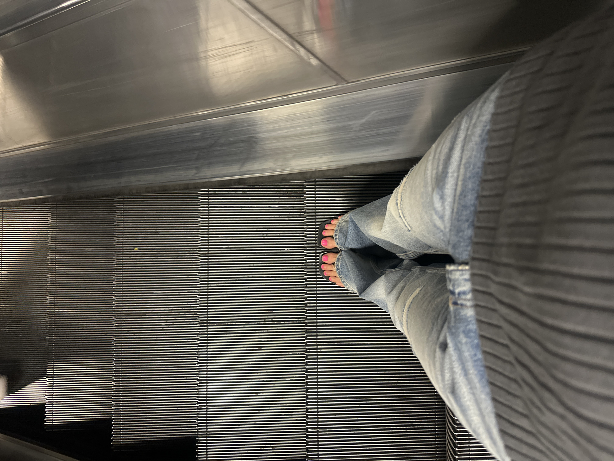 A top-down shot shows a person standing on an escalator. The person is wearing light blue jeans and black sandals with pink toenail polish. The escalator is made of metal with horizontal lines. The escalator handrail is made of stainless steel. The location is likely within a transport hub in London, United Kingdom.