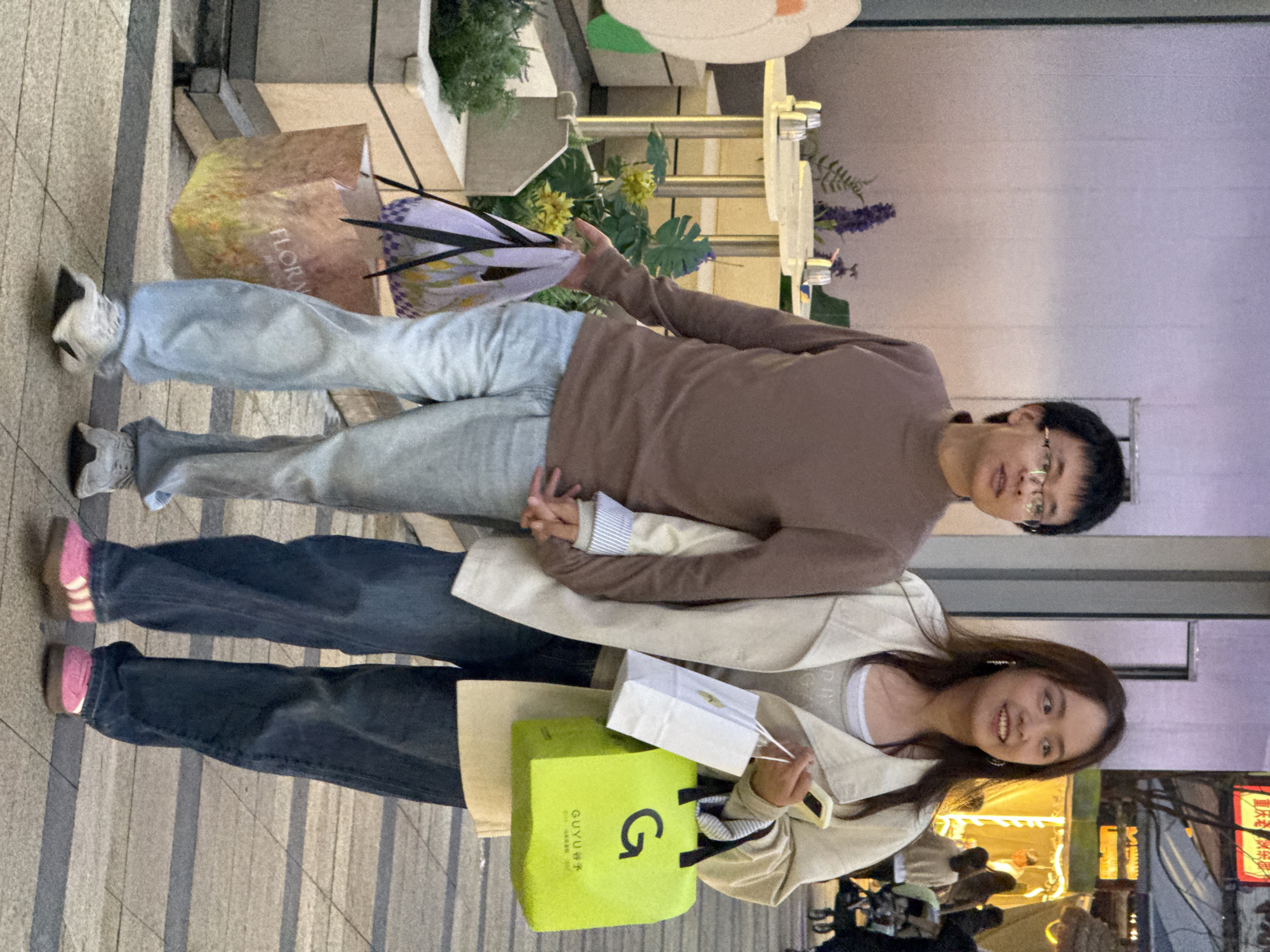 A couple is standing on a sidewalk in Yuzhong District, China. The man is wearing a brown sweater, light blue jeans, and sneakers. He is holding a floral shopping bag and what appears to be a bouquet of flowers. The woman is wearing a beige trench coat, dark blue jeans, and pink socks. She is holding a green shopping bag with a large "G" on it, as well as a small white shopping bag. The street behind them is brightly lit with shops.