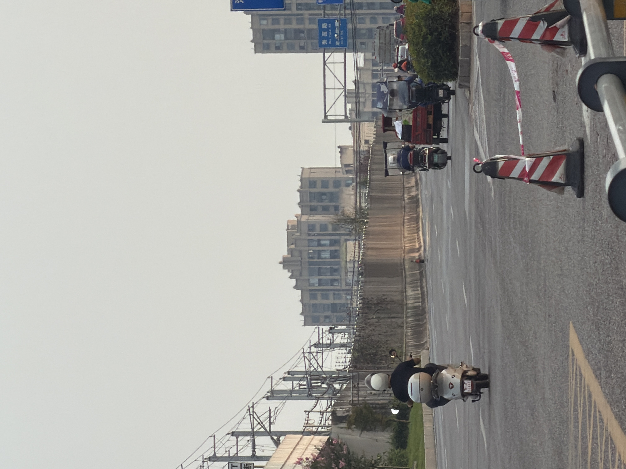 This is a daytime, eye-level shot of a street scene in the city of Jiangjin, China. In the foreground, there are two traffic cones connected by red and white striped tape, indicating a temporary road closure or hazard. The road surface is asphalt with painted white and yellow lane markings.
A person wearing a black shirt and a white helmet is riding a silver scooter away from the camera. The scooter has a large silver storage box mounted on the back and a license plate that reads "56448".
Further down the road, several other vehicles are visible, including three-wheeled motorbikes carrying passengers and cargo, and a truck. A cluster of green bushes is located on the right side of the road.
In the background, a long, textured concrete wall runs horizontally across the image. Beyond the wall, there are several multi-story buildings with numerous windows. Overhead, a complex network of power lines and poles is suspended in the sky, suggesting proximity to a railway or industrial area. The sky is hazy and pale, indicating overcast conditions or light pollution.