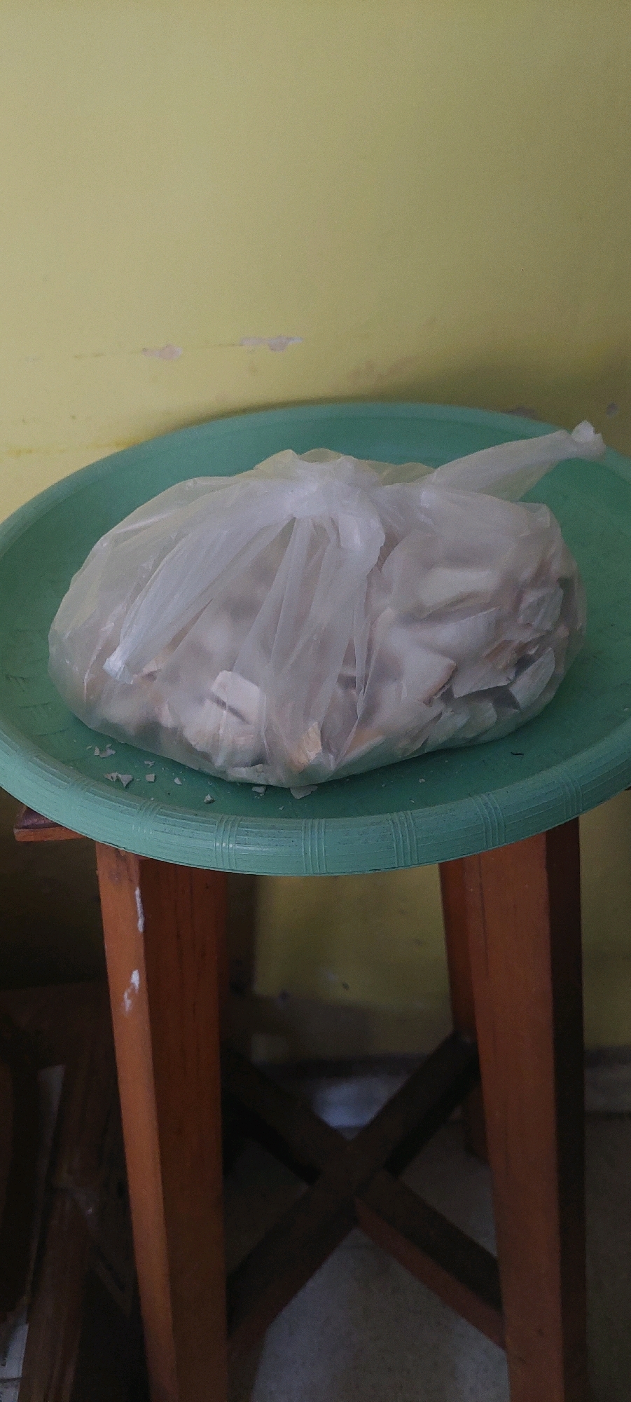 A plastic bag filled with what appears to be dried fish or small bones rests on a green plastic plate. The plate is situated on a wooden stool, the legs of which are visible. The background is a light yellow wall with some peeling paint. The scene is indoors. There are no people visible in the image. The lighting suggests it might be daytime. There are no specific landmarks that would indicate a location in Lumajang, Indonesia.