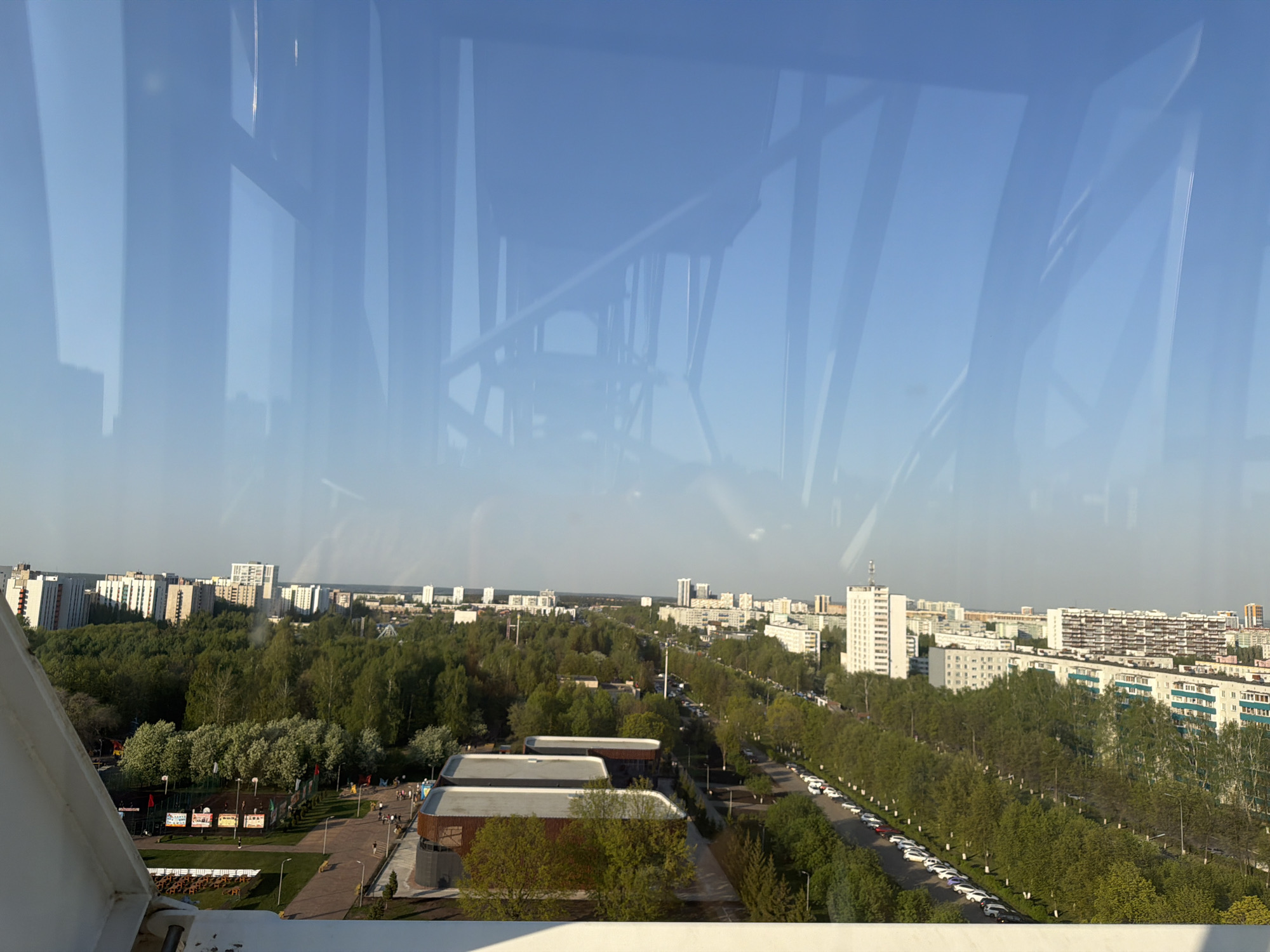 The image captures an elevated view of Naberezhnye Chelny, Russia, likely from a tall structure, possibly a Ferris wheel or similar observation point. The foreground shows parts of the structure on which the photographer is located. The midground presents a cityscape characterized by dense green foliage interwoven with urban infrastructure. A straight tree-lined street runs across the scene. Buildings of varying heights are visible, a mix of residential and commercial structures. The background extends to the horizon under a clear blue sky with indistinct reflections off the glass.