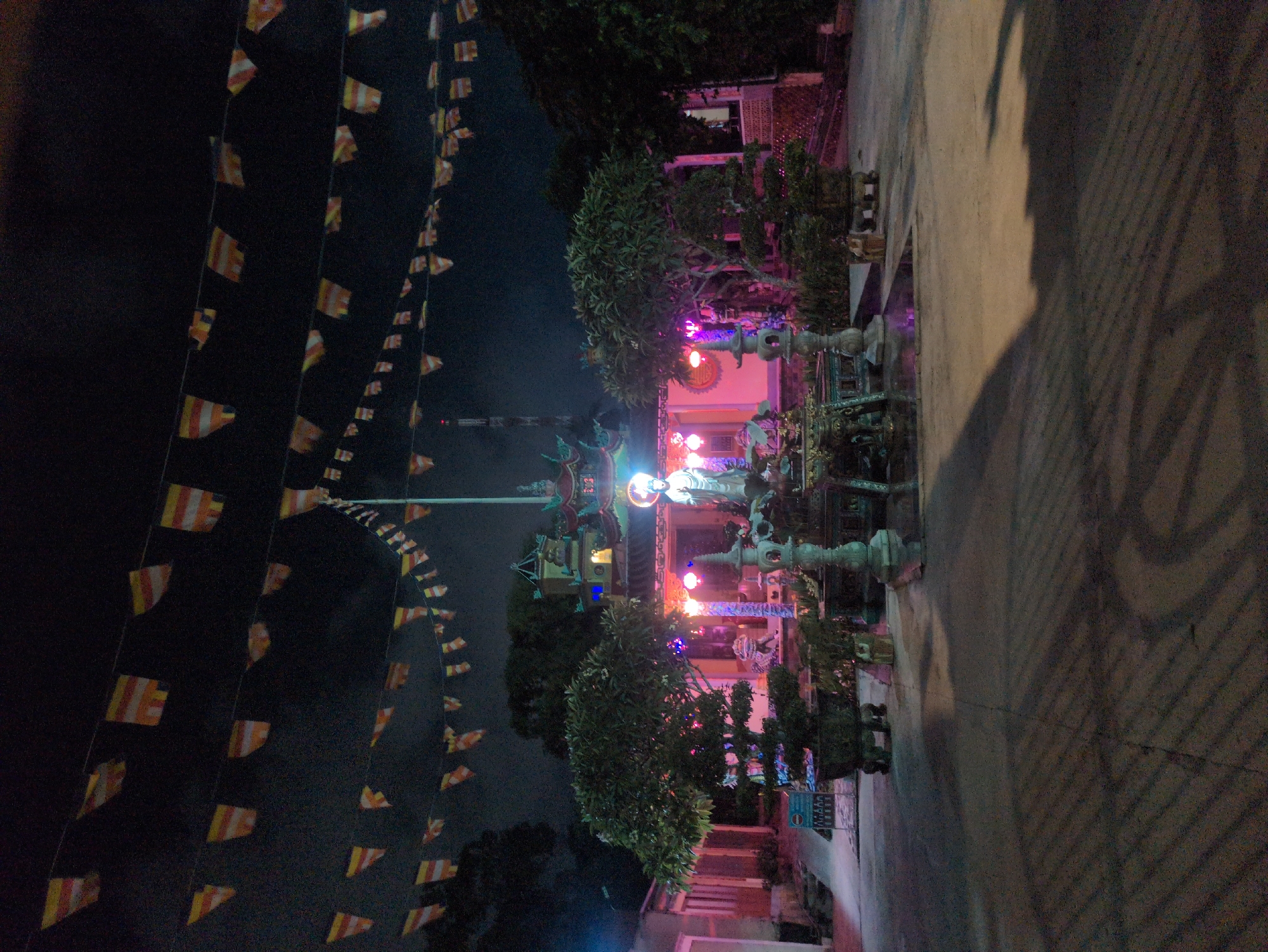 The image depicts a temple at night in Phú Quốc, Vietnam. The temple building is illuminated with pink and white lights, featuring statues on top of the roof and in the courtyard. Strings of Buddhist flags are strung across the foreground, ascending into the dark sky. There are trees and plants around the temple, and a section of paved ground with shadowed areas.