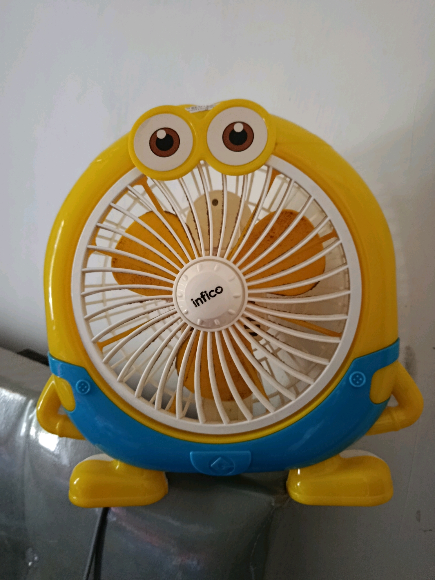 A yellow and blue plastic fan shaped like a Minion character from the Despicable Me franchise is depicted in this medium shot. The fan is positioned centrally against a plain, light-colored wall. The fan's body is primarily yellow, featuring large, cartoonish eyes with brown irises and white circular accents. A blue section wraps around the lower part of the fan, resembling overalls, complete with visible circular blue buttons. The fan blades are visible behind a white grill, and the center of the grill has a white circular component with the black text "infico" on it. The fan has small yellow arm-like protrusions on either side and yellow feet. The overall scene suggests the fan is a decorative and functional item, possibly used in a home environment. The provided context states the image is located in Jambearum, Indonesia, implying this is where the fan is situated. No people or actions are depicted; the image focuses solely on the object.