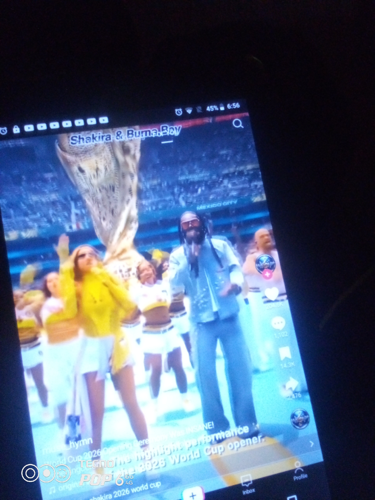 A tablet displays a video featuring a musical performance. In the foreground, a woman in a yellow jacket and white skirt dances. Behind her, other performers are visible. A man in a light blue outfit, wearing sunglasses, stands in the middle ground, also appearing to perform. The background depicts a stadium with a large crowd and a trophy-like structure. Text overlays on the screen read "Shakira & Buma.Boy" and "MEXICO CITY." The video also shows social media engagement metrics like likes and shares. A watermark at the bottom left corner says "TECNOPop 6G 4G." The overall scene suggests a live broadcast or recorded performance of a song related to the World Cup, possibly from Mexico City. There is no indication of Howayi, Nigeria in the image.