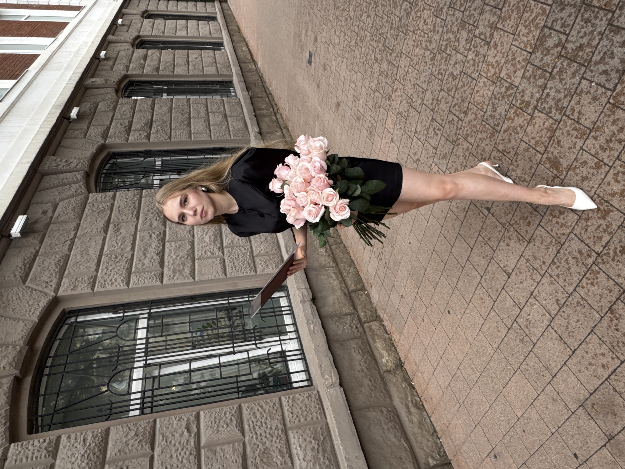 A young woman with blonde hair stands on a sidewalk in front of a stone building. She is wearing a black dress and white high heels. In her right hand, she holds a portfolio or book, and in her left, she carries a large bouquet of pink roses. The building has multiple windows with wrought-iron grates. The sidewalk is paved with reddish-brown tiles. The overall scene suggests an urban environment, possibly in Perm, Russia, given the architectural style and brickwork commonly found in the region. The lighting and wet appearance of the sidewalk indicate overcast or recent rain conditions.