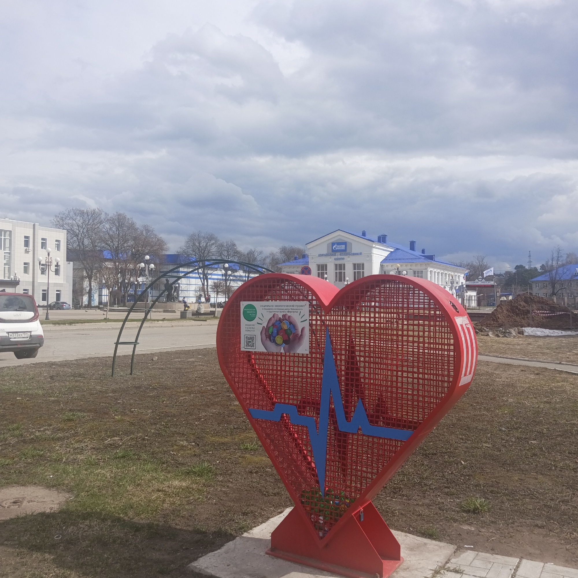The image shows a large, red, heart-shaped structure in the foreground, likely a container for collecting plastic bottle caps, located in Chaikovsky, Russia. A blue ECG waveform is affixed to the structure. A sign on the heart promotes a social-ecological project to support special needs children of Chaikovsky. The container is on a concrete base amidst a patch of sparse grass and dirt. A paved area extends behind the heart, leading to buildings in the background, including one with a Gazprom logo. A grey sky filled with clouds overhangs the scene. A white car is parked along a street adjacent to the scene. An arched metal structure stands behind the heart.