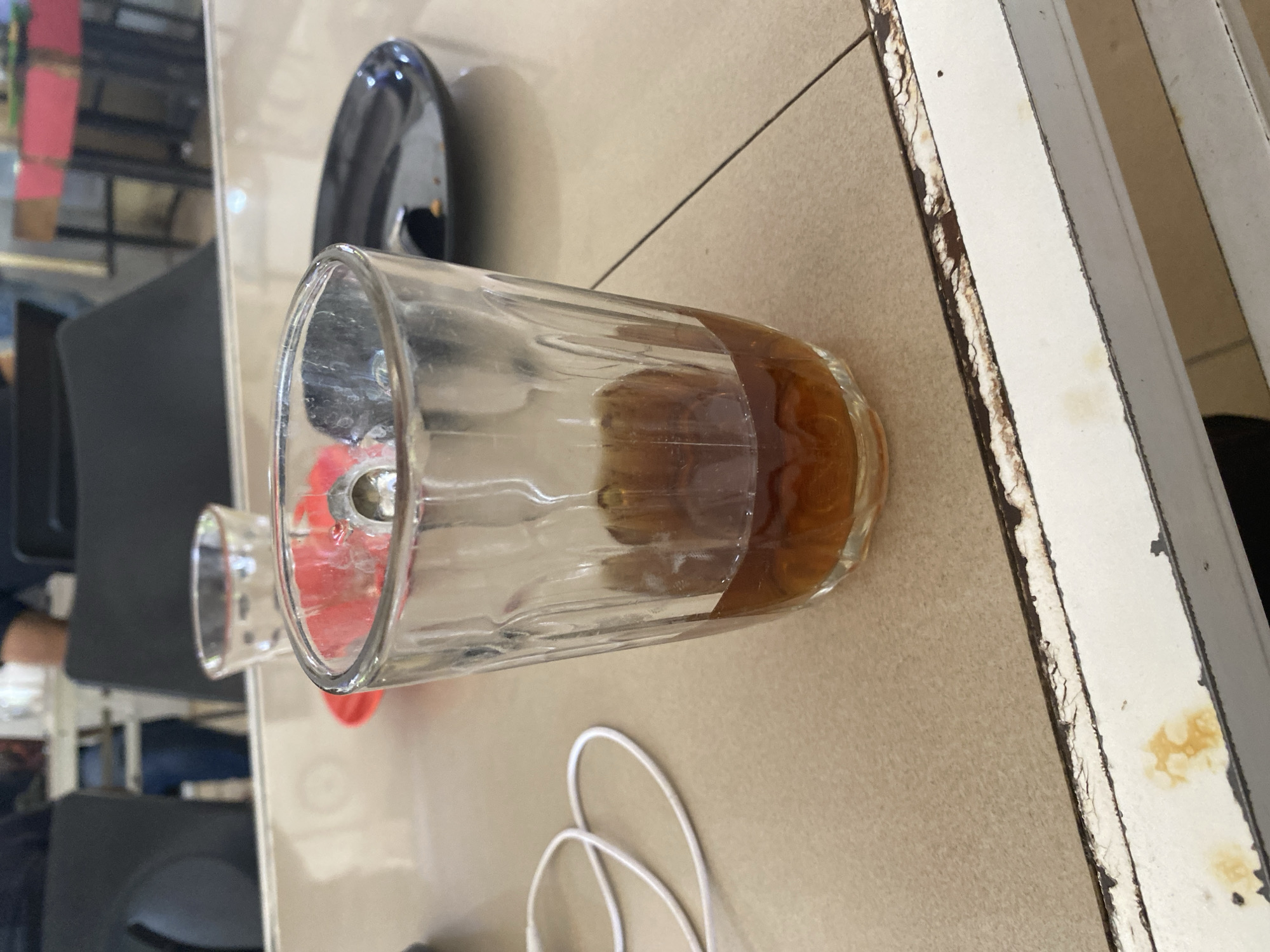 In a close-up shot, a glass half-filled with a dark brown liquid, likely tea or coffee, sits on a tiled surface. The glass is textured with vertical lines and a subtle rippled effect. To the left of the glass, a white cord, possibly from earbuds or a charger, is loosely coiled. In the background, out of focus, are glimpses of a restaurant or cafe setting. Two dark chairs are visible, and further back, a red and white structure suggests a serving counter or food stall. The overall scene is of a casual dining environment. The image was taken in Lhokseumawe, Indonesia, at a location likely a cafe or restaurant.