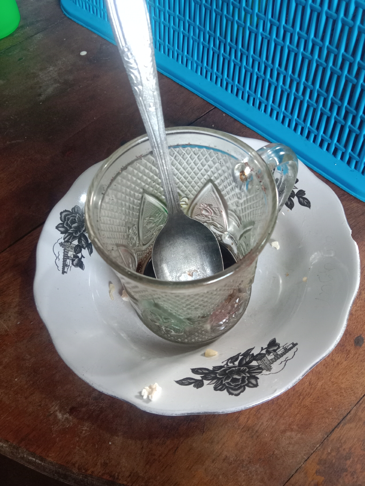 A close-up, overhead shot shows a glass coffee cup with a spoon inside, placed on a saucer. The cup and saucer are white with black floral and building motifs. Some white food residue is visible on the saucer and the wooden table it rests on. In the background, a blue plastic basket is partially visible. The scene appears to be indoors, on a wooden table, possibly in a home or small eatery in Paya Besar, Indonesia.
