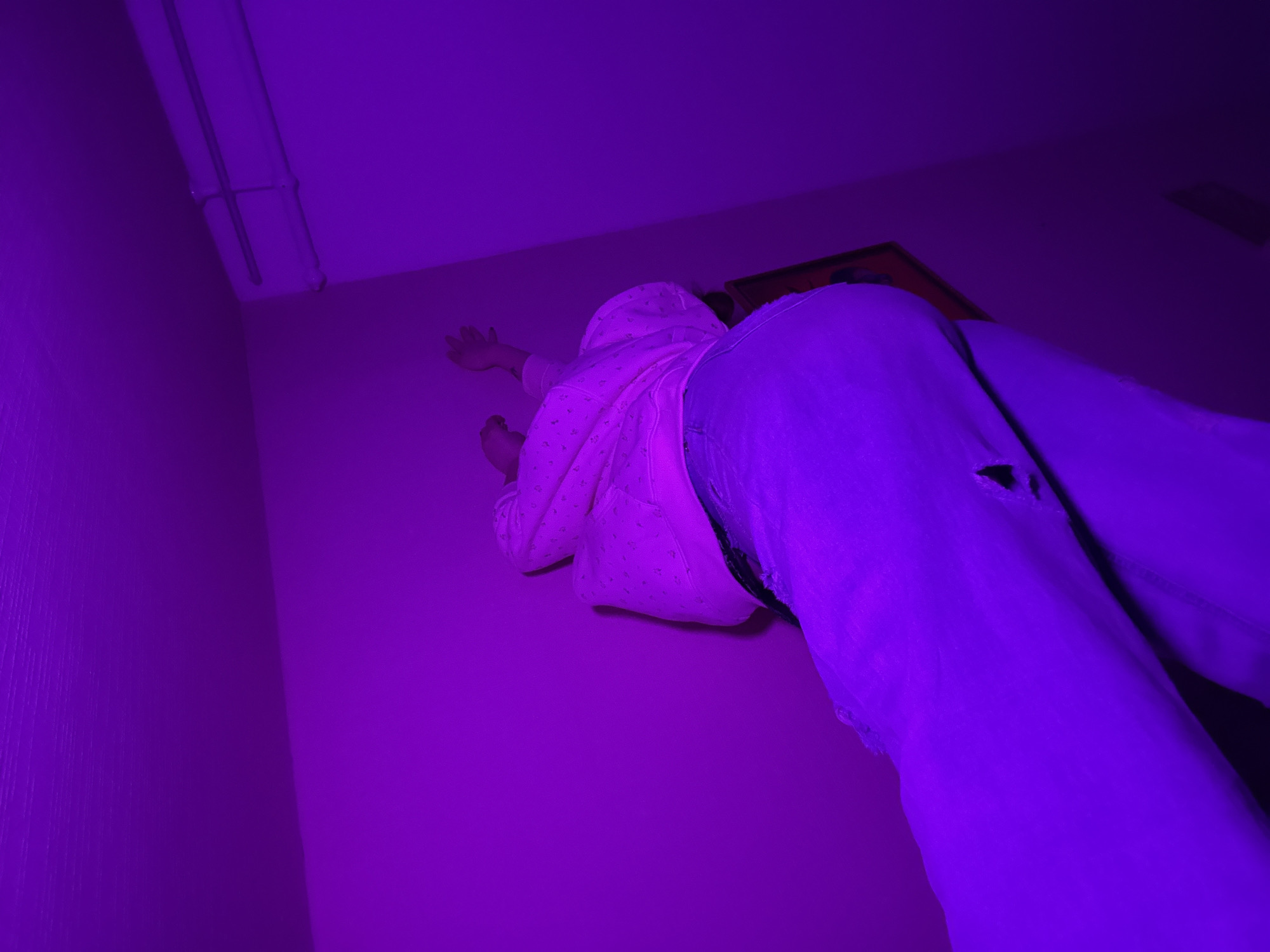 The image depicts a person lying against a wall. The person is wearing jeans with a hole, and a hooded jacket. The lighting is purple. The location is inside a room with a fluorescent light fixture and an object on the wall. There is no verifiable information to suggest the location is in Izhevsk, Russia.