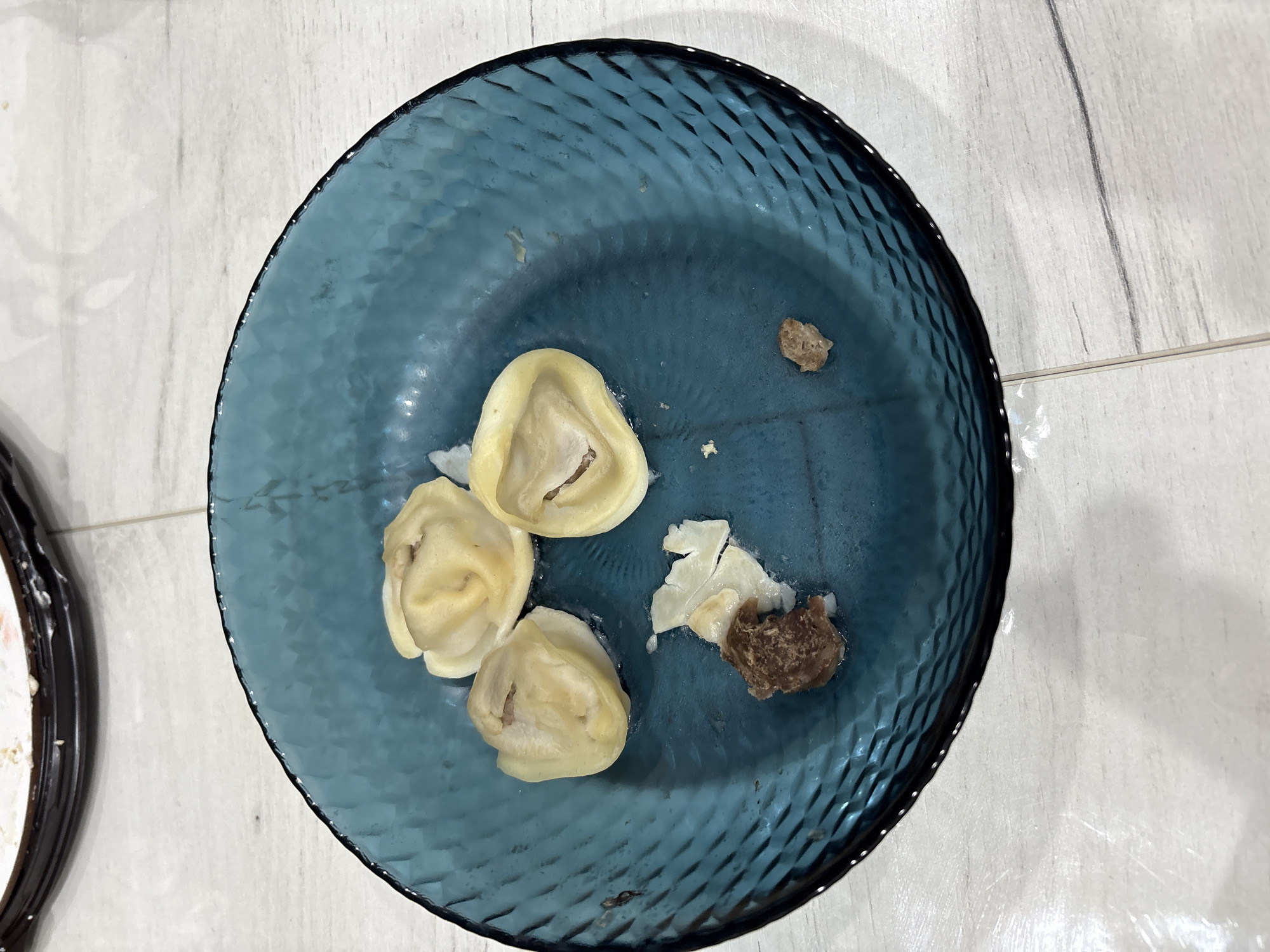 A blue, textured glass plate rests on a light-colored, tiled surface. The plate contains three cooked, stuffed pasta dumplings, and a torn piece of pasta with a bit of meat filling. There is also a small, single piece of meat filling. The location is unknown.