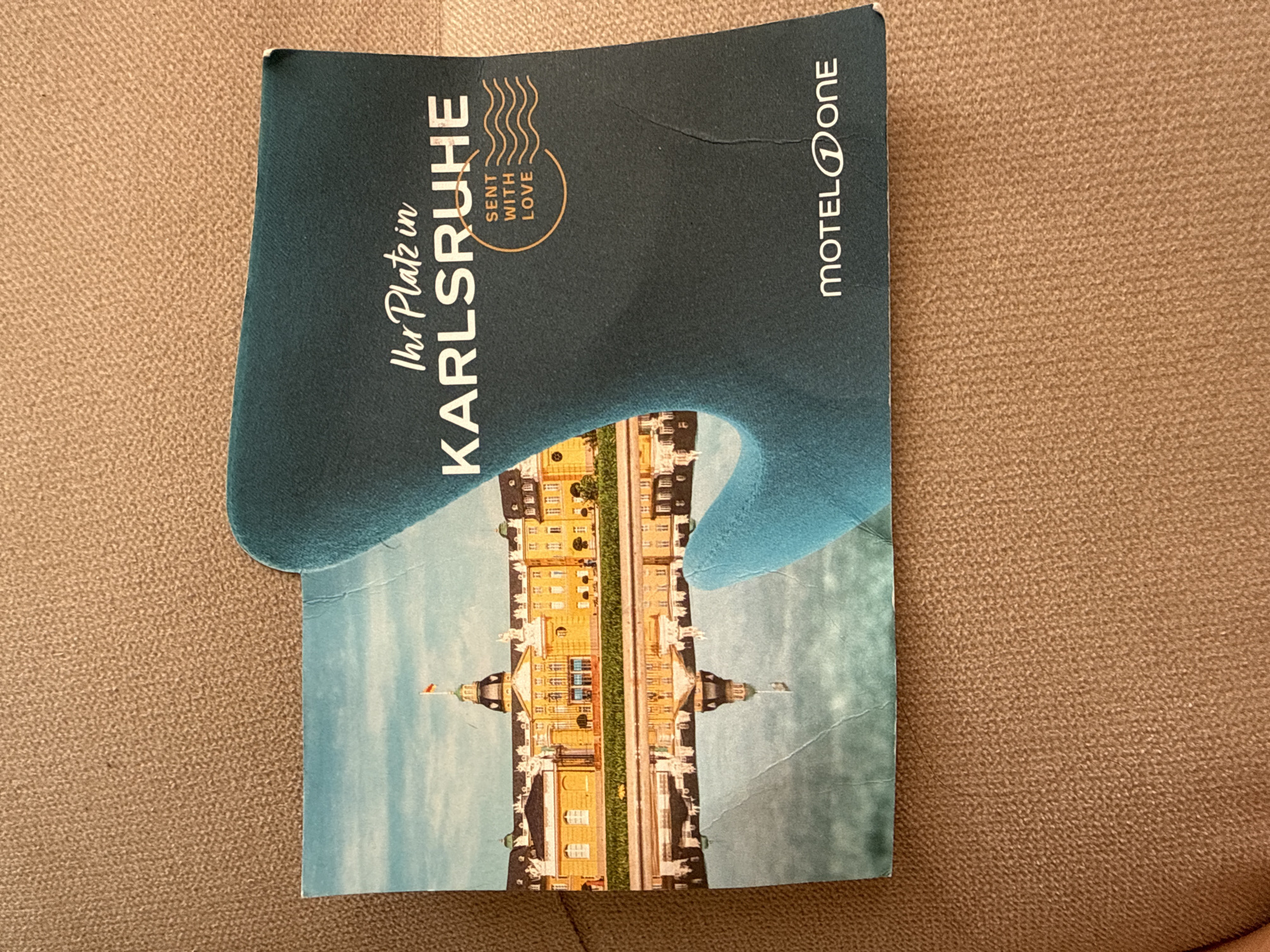 A folded promotional brochure or postcard, predominantly dark teal and light blue, rests on a textured, light brown surface resembling carpet or upholstery. The left panel of the brochure displays a photograph of a large yellow palace building with a central domed tower topped with a small red and white flag. The palace is reflected clearly in calm water in the foreground, with a strip of green grass separating the building from the water. The sky above the palace is light blue. The right panel, colored dark teal, features white text. The top text reads "Ihr Platz in" in a script font, followed by "KARLSRUHE" in a bold, sans-serif, all-caps font. Below this, a circular graphic in light brown outlines the text "SENT WITH LOVE" in white, above three horizontal wavy lines, also in light brown. At the bottom right of the dark teal panel, the white "MOTEL ONE" logo is visible. The brochure exhibits several creases, particularly on its left side and along the fold line. The scene is evenly lit from above. The location indicated on the brochure is Karlsruhe.