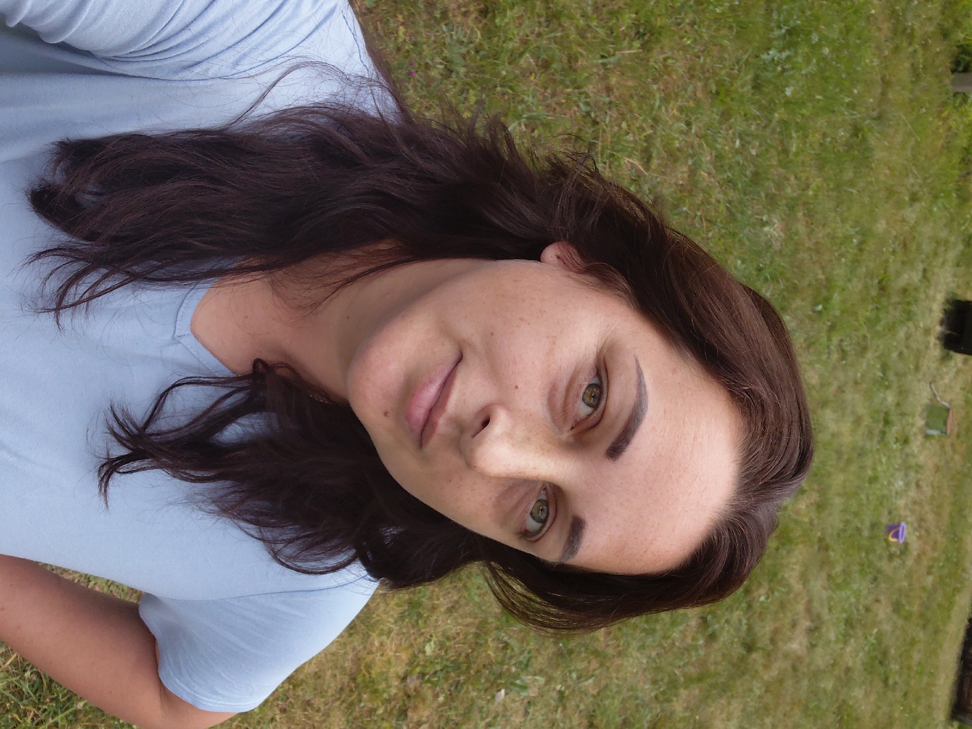 A woman with long, dark brown hair and green eyes, wearing a light blue t-shirt, poses for a selfie outdoors in a grassy area. Her hair is wavy and falls over her shoulders. She has a light dusting of freckles across her nose and cheeks. Her left hand is resting on her hip. In the background, out of focus, is a green lawn with some scattered small objects, possibly toys, and what appear to be fence posts. The scene suggests a casual outdoor setting, possibly a backyard or park.