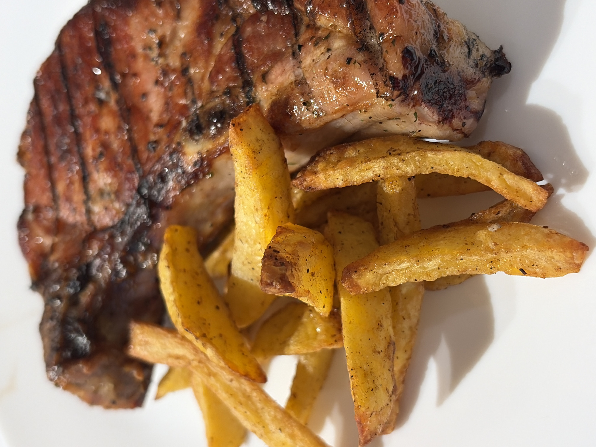 The image depicts a pork chop, grilled with visible grill marks, served alongside a pile of french fries. The food is presented on a white plate, likely indoors under bright lighting. There's no information to suggest the location is Chaikovsky, Russia, or any details involving people, specific actions, or interactions. The focus is solely on the plated food.