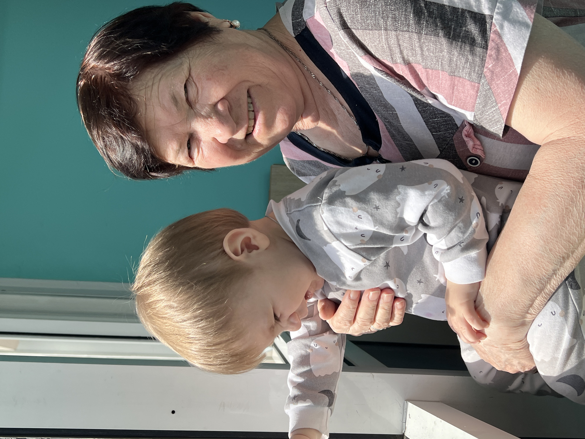 A heartwarming moment between an older woman and a young child is captured in this image from Perm, Russia. The woman, with short dark brown hair, is looking directly at the viewer with a broad, joyful smile. Her face is illuminated by bright sunlight, highlighting her features and expressive eyes. She is wearing a horizontally striped shirt in shades of pink, grey, white, and black, along with a silver-colored necklace and a pearl earring. Her hands are gently holding the child.
The young child, with light blonde hair, is dressed in a grey and white patterned jumpsuit featuring cartoon sheep or llamas, stars, and crescent moons. The child's attention is directed downwards, looking out of frame, while being held securely by the woman.
The scene is bathed in strong, direct sunlight, indicating a clear, bright day, likely morning or late afternoon given the distinct shadows. A vibrant turquoise or teal wall forms a significant part of the background on the left, suggesting an accent wall indoors or a painted exterior. A white railing or structure is visible at the bottom of the frame, potentially a window frame or a balcony barrier. The image conveys a sense of warmth, happiness, and familial affection. There is no visible text.