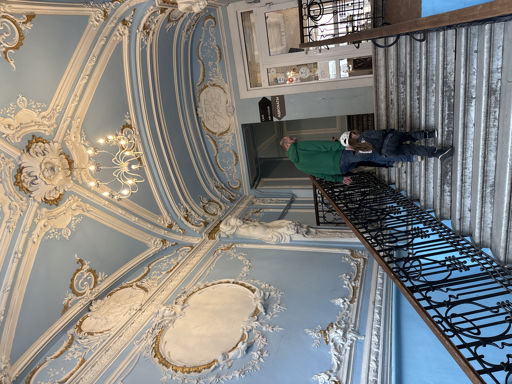 The image depicts the interior of a building in Saint Petersburg, Russia, characterized by ornate Baroque-style architecture. The walls and ceiling are light blue with white and gold decorative plasterwork, including moldings, floral patterns, and figurative reliefs. A chandelier hangs from the ceiling. In the foreground, a black wrought-iron staircase ascends. Two people are visible on the stairs: one wearing a green jacket and the other wearing black and white hat and clothing. A sign indicates "Cafe Art Cloud" and "Advocates." Above the stairs, a doorway leads to the exterior, with a window displaying signage in Russian. A metal roll-up door partially covers the entrance.