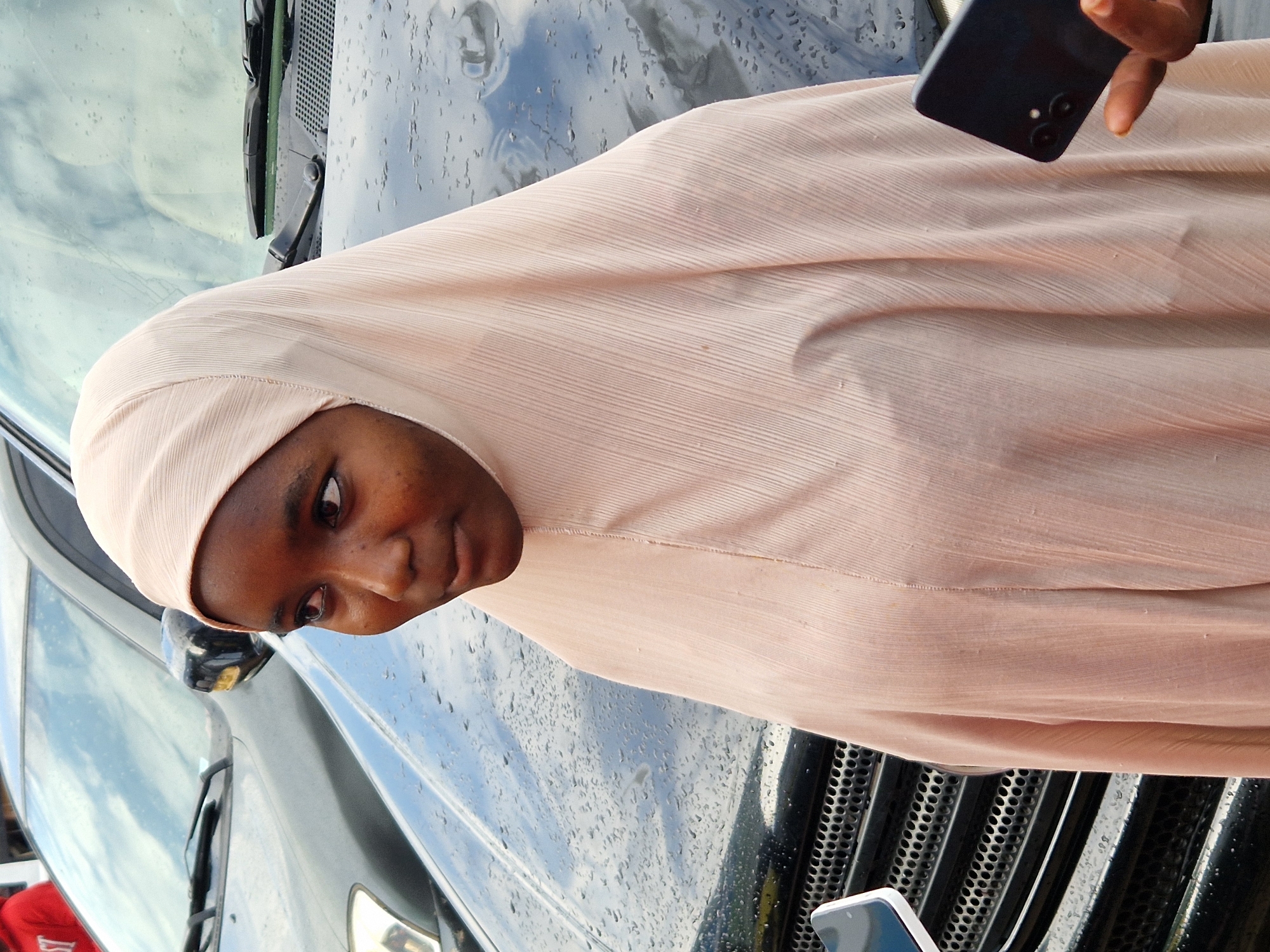 A young Black woman wearing a light pink hijab and clothing stands in front of a dark-colored car with water droplets on its hood. She is looking to her left. Her right hand is holding a dark-colored smartphone. A portion of another smartphone is visible in the lower left corner of the image. The background shows the reflection of the sky on the car's surface.
