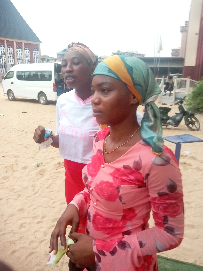 Two young women stand in a sandy area. The woman in the foreground wears a floral pink shirt and a headscarf. She also has a gold necklace. The woman behind her wears a white t-shirt and a red skirt. She also wears a headscarf. A white van is parked in the background. A motorcycle and building are also visible.