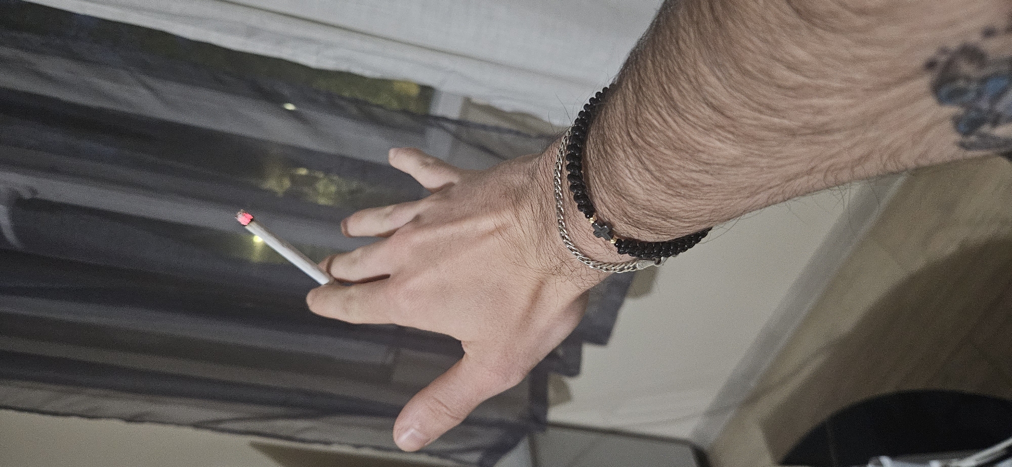 A male hand is holding a lit cigarette against a background of light-colored window blinds and black mesh. The person has a beaded bracelet and a silver chain bracelet on their wrist, and a tattoo is visible on their upper arm. The location is likely indoors and not specifically identifiable within the city of Plovdiv, Bulgaria, based on the visual information provided.