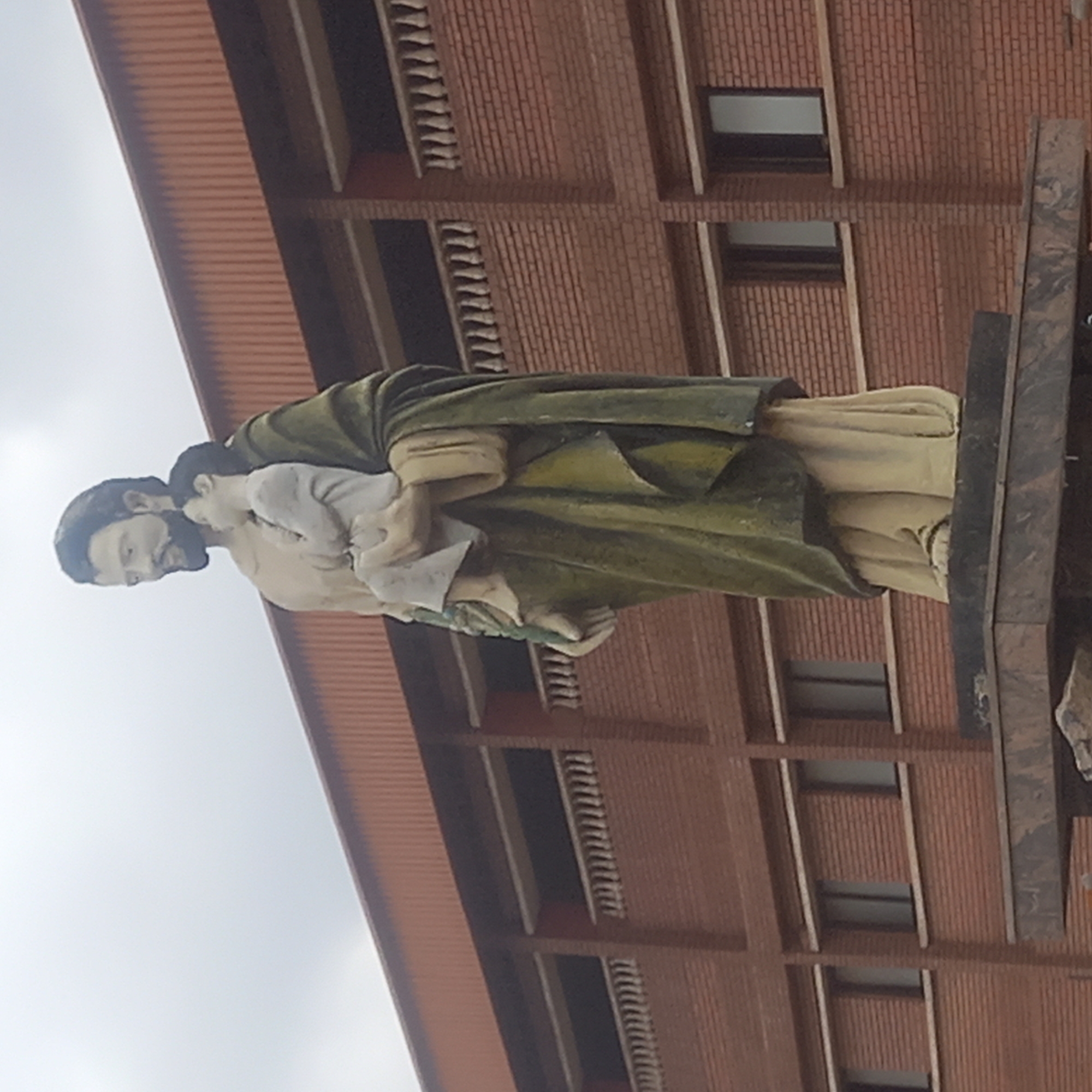 The image shows a statue of Saint Joseph carrying the child Jesus, situated in Sardauna, Nigeria. The statue, composed of light-colored figures with dark green accents, is mounted on a dark grey or black rectangular base. The background features a multi-story brick building with regularly spaced windows and a red corrugated roof. The sky is visible at the top left corner, appearing overcast.