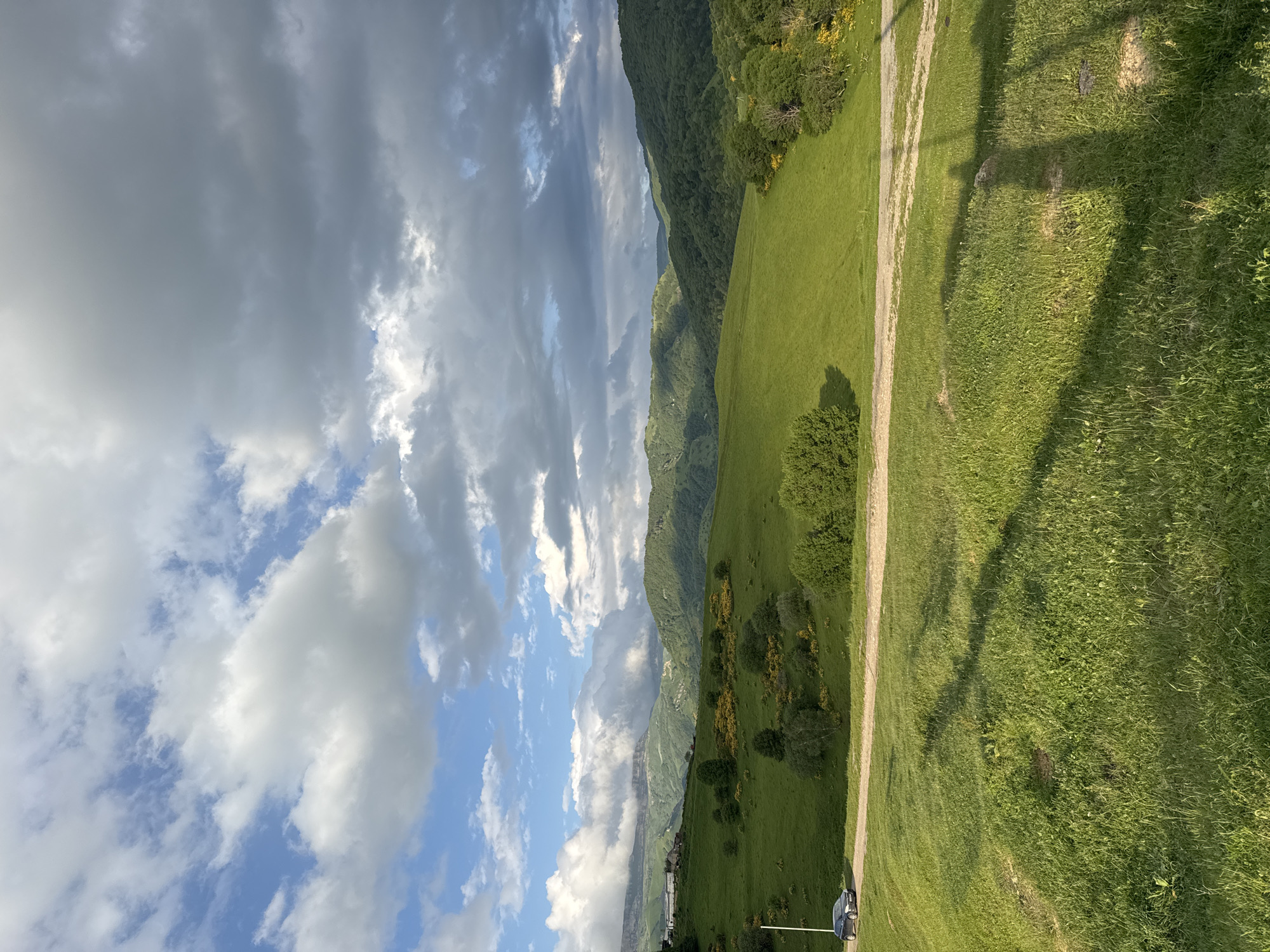 The image depicts a vast, green, rolling landscape under a dramatic sky filled with both white and dark grey clouds. In the foreground, a grassy slope transitions into a dirt road that curves through the middle ground. A dark-colored car is parked on the left side of the road. The mid-ground features more lush green hills dotted with trees and patches of yellow flowering bushes. In the background, a series of layered, tree-covered mountains recede into the distance, with some of the peaks partially obscured by clouds. The lighting suggests it is late afternoon or early evening, with shadows cast across the foreground grass. The scene is one of natural beauty and apparent tranquility, likely in a rural or mountainous region such as the area around Nizhnyaya Mara, Russia.