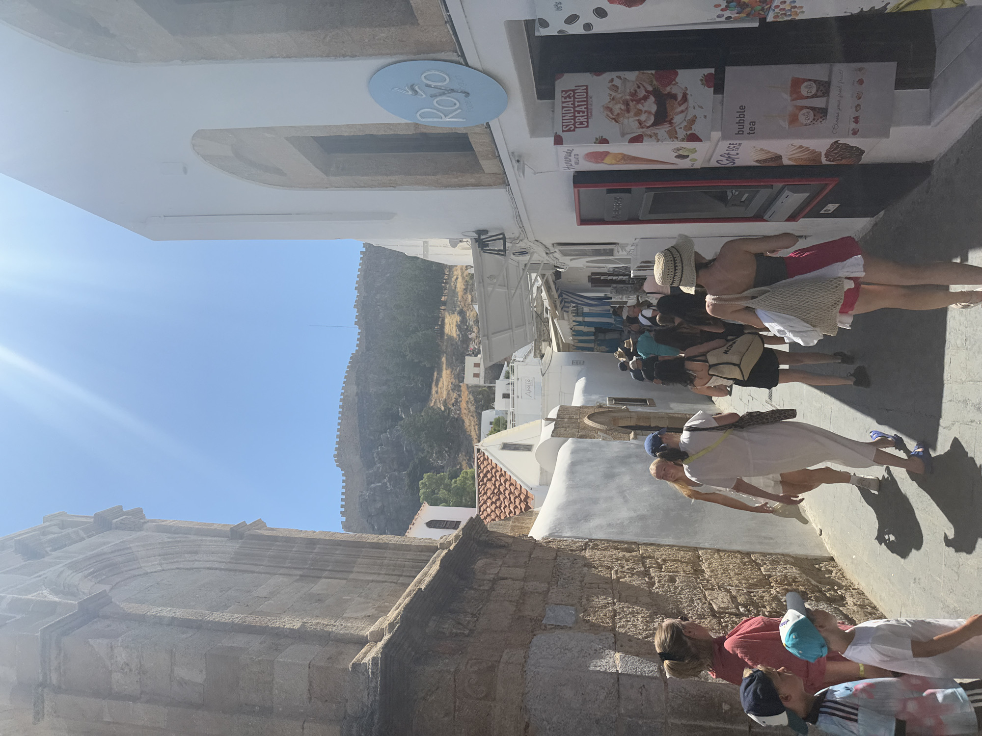 A group of people are walking down a narrow, sun-drenched cobblestone street in Lindos, Greece. The street is flanked by whitewashed buildings with traditional architecture. On the left, a large stone building with arched openings dominates the foreground. On the right, a building with a bright blue circular sign that reads "Rovo" features an advertisement for "Sundaes Creation" and "Bubble Tea." Several individuals, including children, are visible, dressed in casual summer attire. The sun casts long shadows on the ground. In the background, the Acropolis of Lindos and the surrounding ancient fortifications are visible on a rocky hilltop under a clear blue sky.