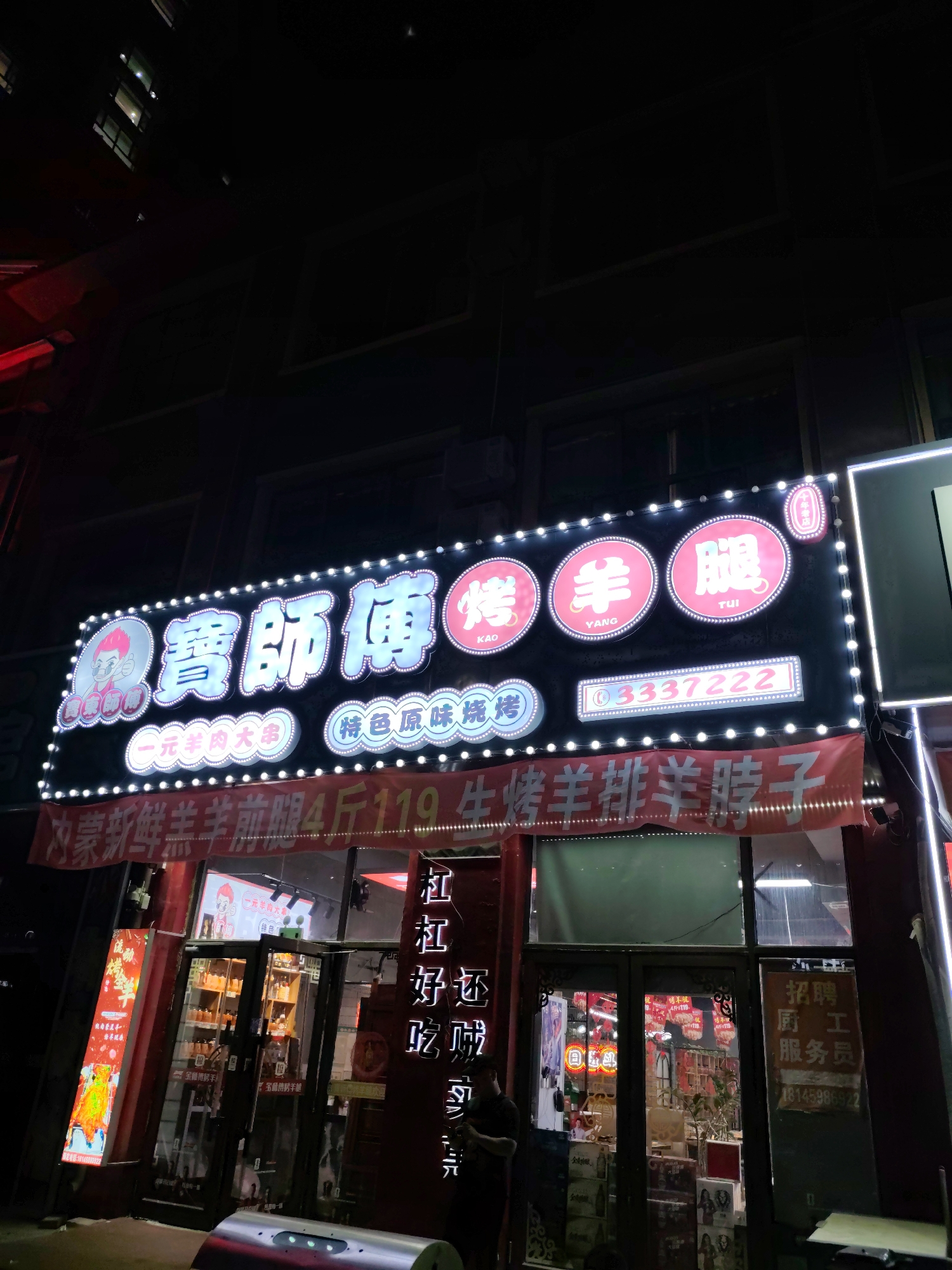 The image shows a brightly lit storefront at night in Lindian, China. The storefront features a large, illuminated sign with Chinese characters and images, indicating a restaurant or food establishment. The sign is adorned with numerous small white lights. Below the main sign, a red banner displays prices and names of food items in Chinese characters. The entrance to the establishment is a glass door, through which shelves with bottles and other items are visible. To the left of the entrance, a vertical illuminated sign displays an image and Chinese text. A person is standing near the entrance, looking at their phone. The environment appears to be an urban street scene at night.