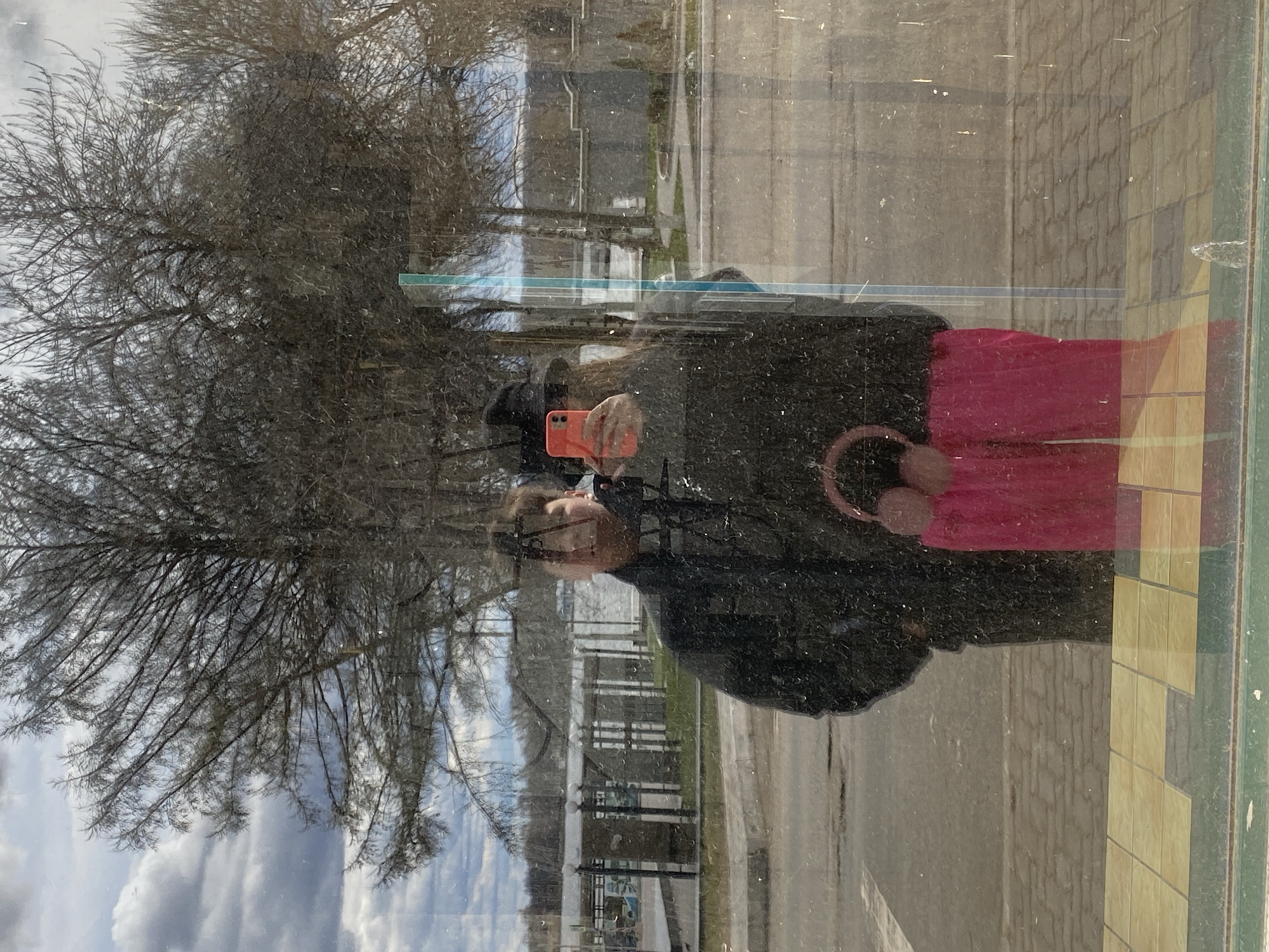 This image captures a selfie reflected in a grimy, reflective surface, likely a large window, in what appears to be an urban or suburban setting in Votkinsk, Russia. The perspective is from the individual taking the photo.
The primary subject is a person, visible in reflection, holding a bright orange smartphone to take the picture. They are wearing a dark, possibly padded, jacket or coat and bright pink pants. A pair of pink over-ear headphones rests around their neck or is held in their other hand. Their face is partially obscured by the phone, but glasses are discernible.
The reflected background behind the person shows an outdoor scene under a cloudy sky. Numerous bare, deciduous trees, devoid of leaves, dominate the left and upper portions of the reflection, suggesting it is late autumn, winter, or early spring. Beyond the trees, there are indistinct buildings or structures, and what appears to be a road or paved area.
The reflective surface itself shows visible streaks, smudges, and dust particles, indicating it is an outdoor window or similar panel. On the far right edge of the image, the exterior of the building where the reflection is taking place is partially visible, featuring a pattern of yellow and green rectangular tiles.
The activity taking place is a self-portrait (selfie) captured through a reflection. The time of day is daytime, with natural light illuminating the scene, albeit under overcast conditions. No discernible text is visible on the phone screen or elsewhere in the image.