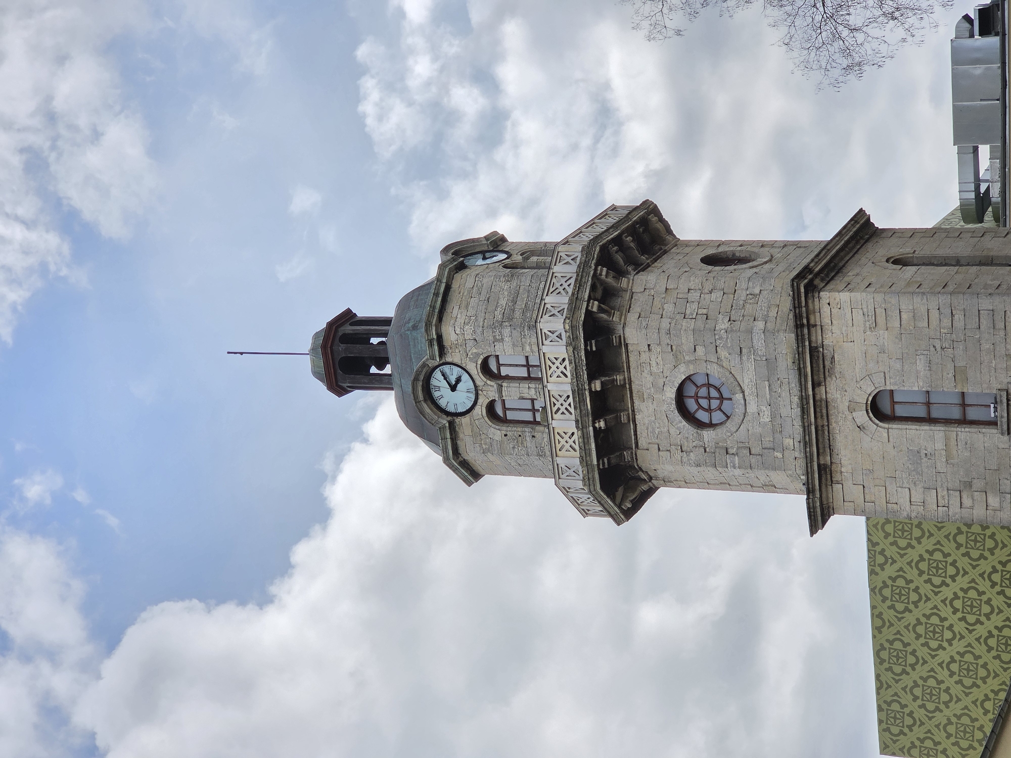 A low-angle shot shows a stone clock tower with a dark green dome and weathervane set against a cloudy blue sky. The clock tower features a large clock face, arched windows, and decorative stonework, as well as visible damage or wear. A building abuts the tower and features a green patterned decoration. The clock tower is the Varna Clock Tower, which is located in Varna, Bulgaria.