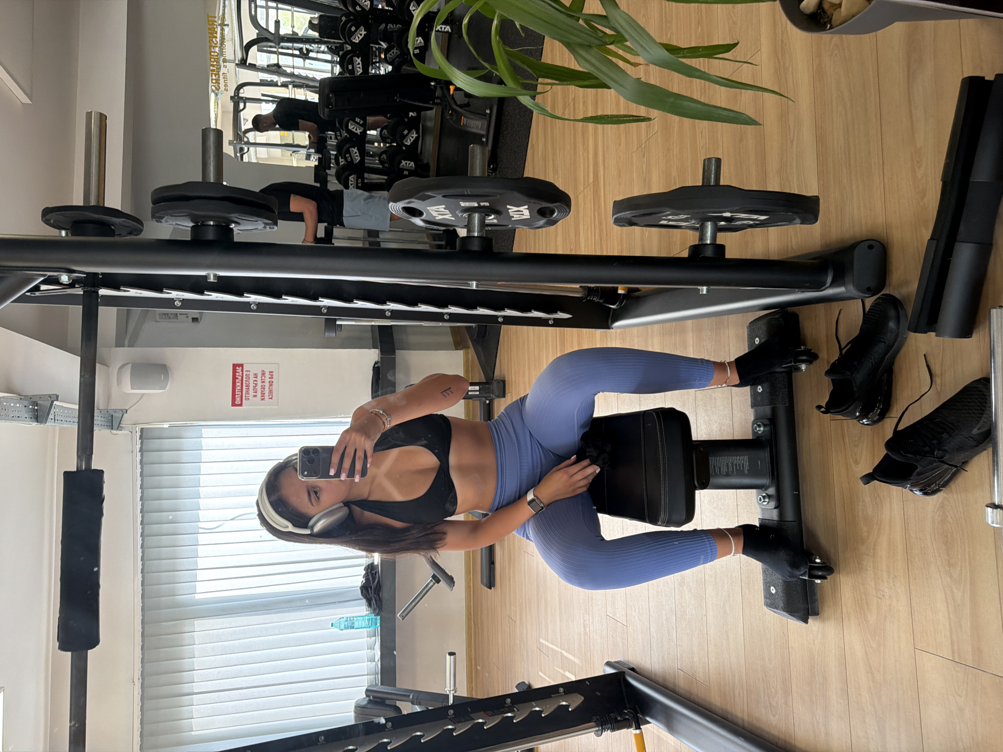 A young woman with dark hair and headphones is sitting on a gym bench, taking a selfie in the mirror. She is wearing a black sports bra and light blue leggings. To her left, there is a weightlifting rack with barbells and weights. In the background, other gym equipment is visible, including weight racks and mirrors. The floor is made of light-colored wood. The overall scene depicts a gym environment. The location is identified as a gym in Gabrovo, Bulgaria, based on the text on the poster in the background which reads "ТРАНСФОРМЕРИЗЪМ 2 FITNESS", "gabrovo.bg" and "8494".