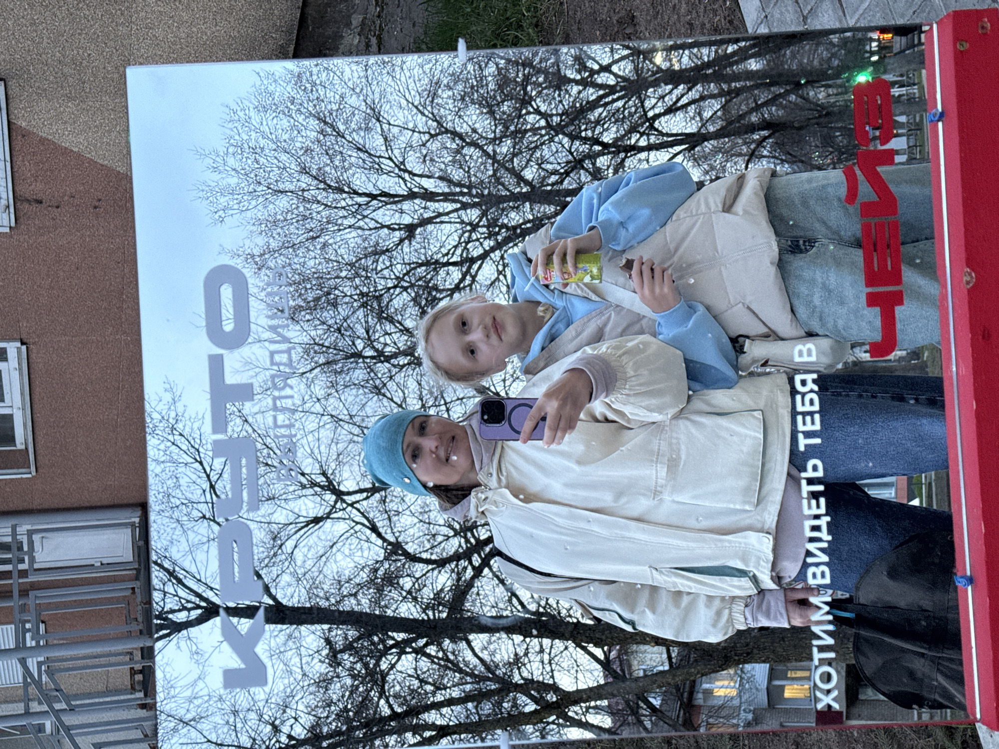 Two individuals, one adult female and one child, are reflected in a mirror. The adult, wearing a light-colored jacket and a blue beanie, is holding a purple smartphone, presumably taking a selfie. The child, wearing a light blue hoodie, is holding a juice box.
The background reflected in the mirror shows bare trees, buildings, and a sky suggesting early evening or overcast conditions. The mirror itself has the text "КТО ХОТИМ ВИДЕТЬ ТЕБЯ" and "СВЕТ" written on it.
The location is likely Chaikovsky, Russia, as indicated by the writing on the mirror. The surrounding environment appears to be urban, with residential buildings visible.