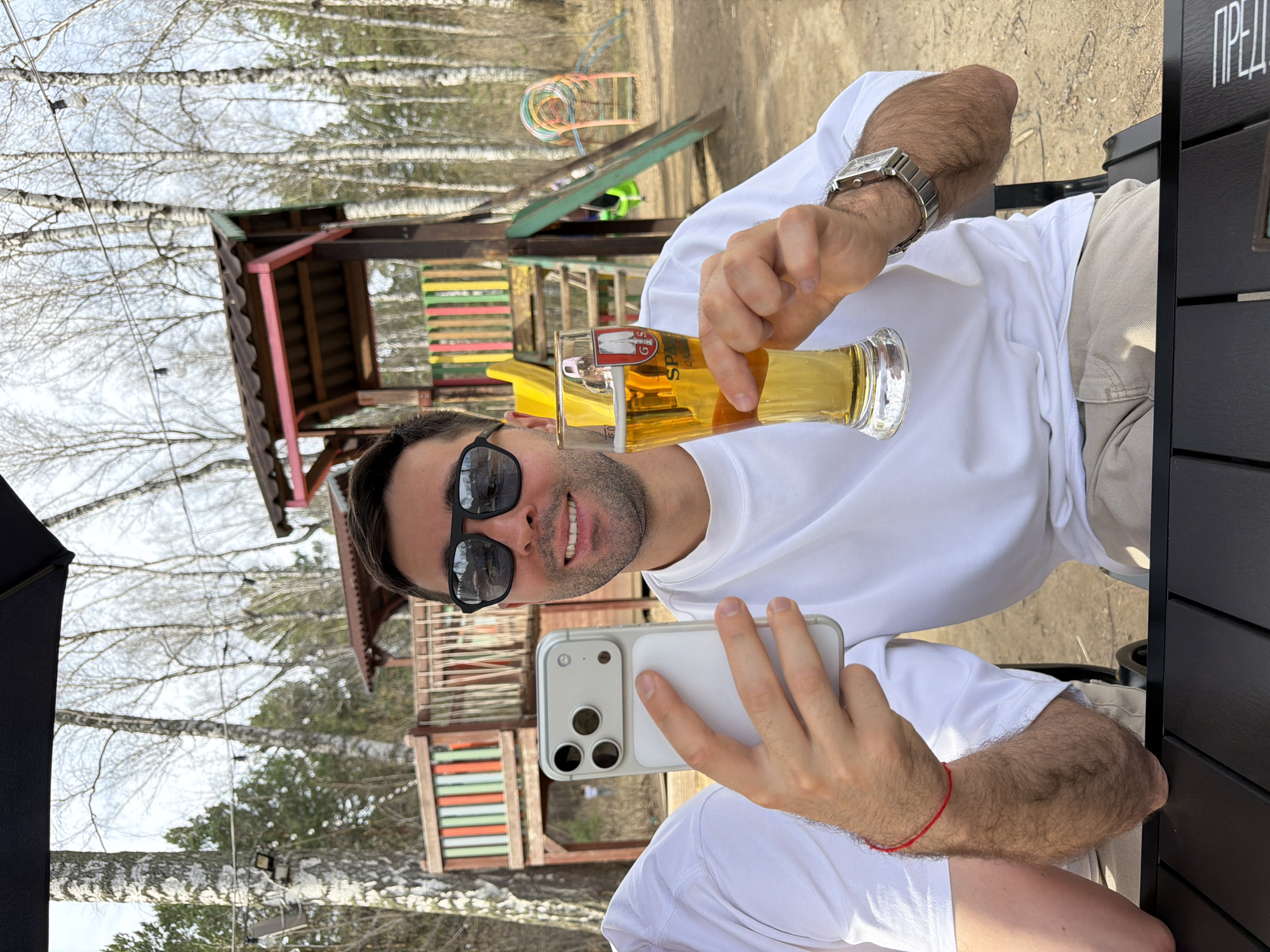 A man is sitting at an outdoor table, taking a photo of himself holding a glass of beer. He is wearing a white t-shirt, khaki pants, and sunglasses, and a metal wristwatch on his left wrist. His phone is a white iPhone with three camera lenses. The beer glass is branded "SPAR." Behind him is a playground with a wooden play structure and slide. The environment suggests a park or recreational area in Krasnoyarsk, Russia.