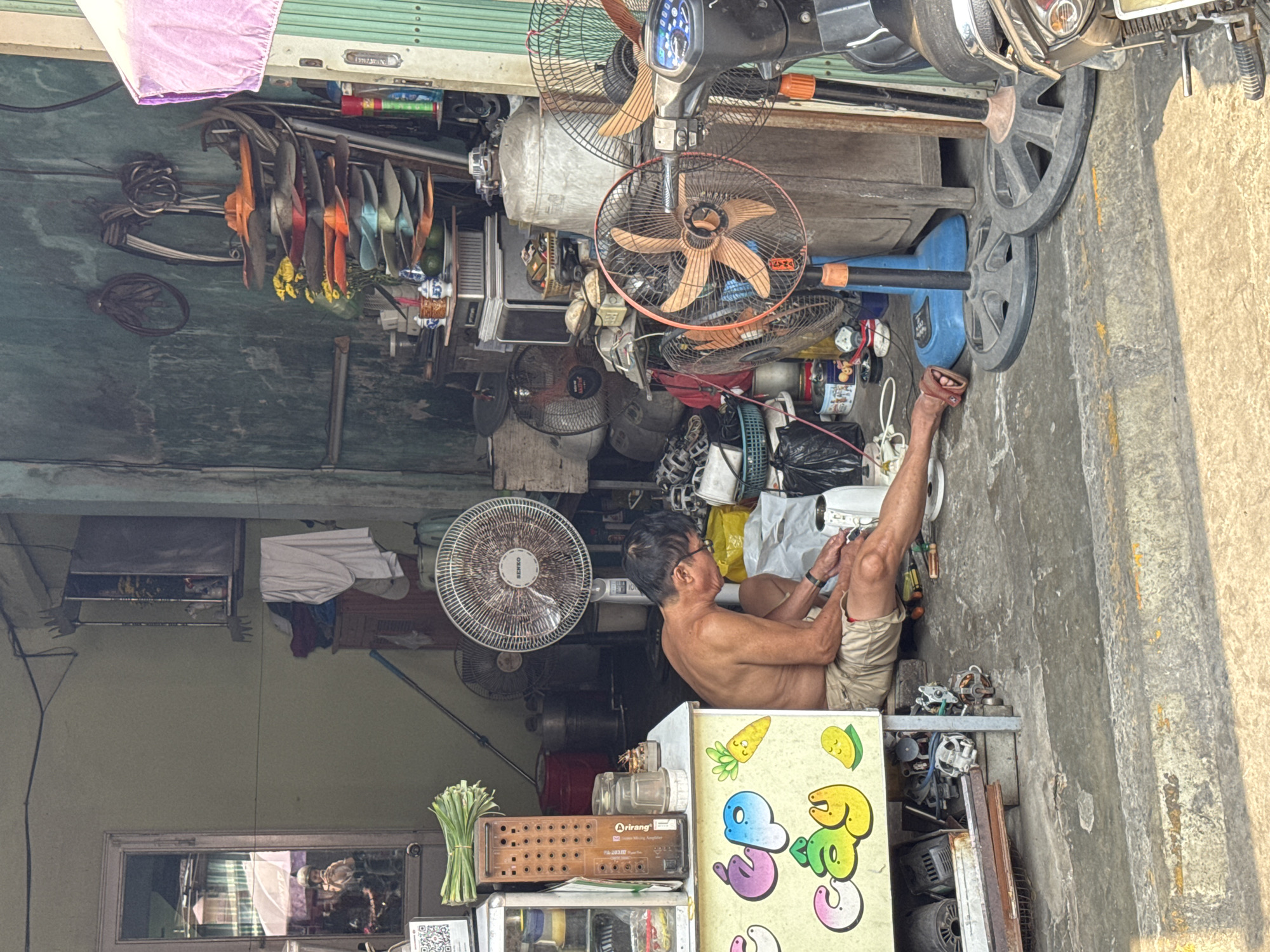 The image depicts an elderly man in a cluttered storefront in Đà Nẵng, Vietnam. He sits shirtless on the floor, wearing khaki shorts and sandals, his legs crossed. He is engrossed in working with small tools in his hands, suggesting he is repairing something.
The environment is densely packed with various objects, including numerous electric fans, both standing and disassembled, indicating a potential repair shop. A motorcycle is parked partially inside the store, adding to the cramped feeling.
Various items, such as spare parts, tools, and electrical components, are scattered around the man, illustrating the disarray of the workshop. The backdrop shows a faded building structure. The image conveys a sense of a small, informal business in a bustling urban environment.