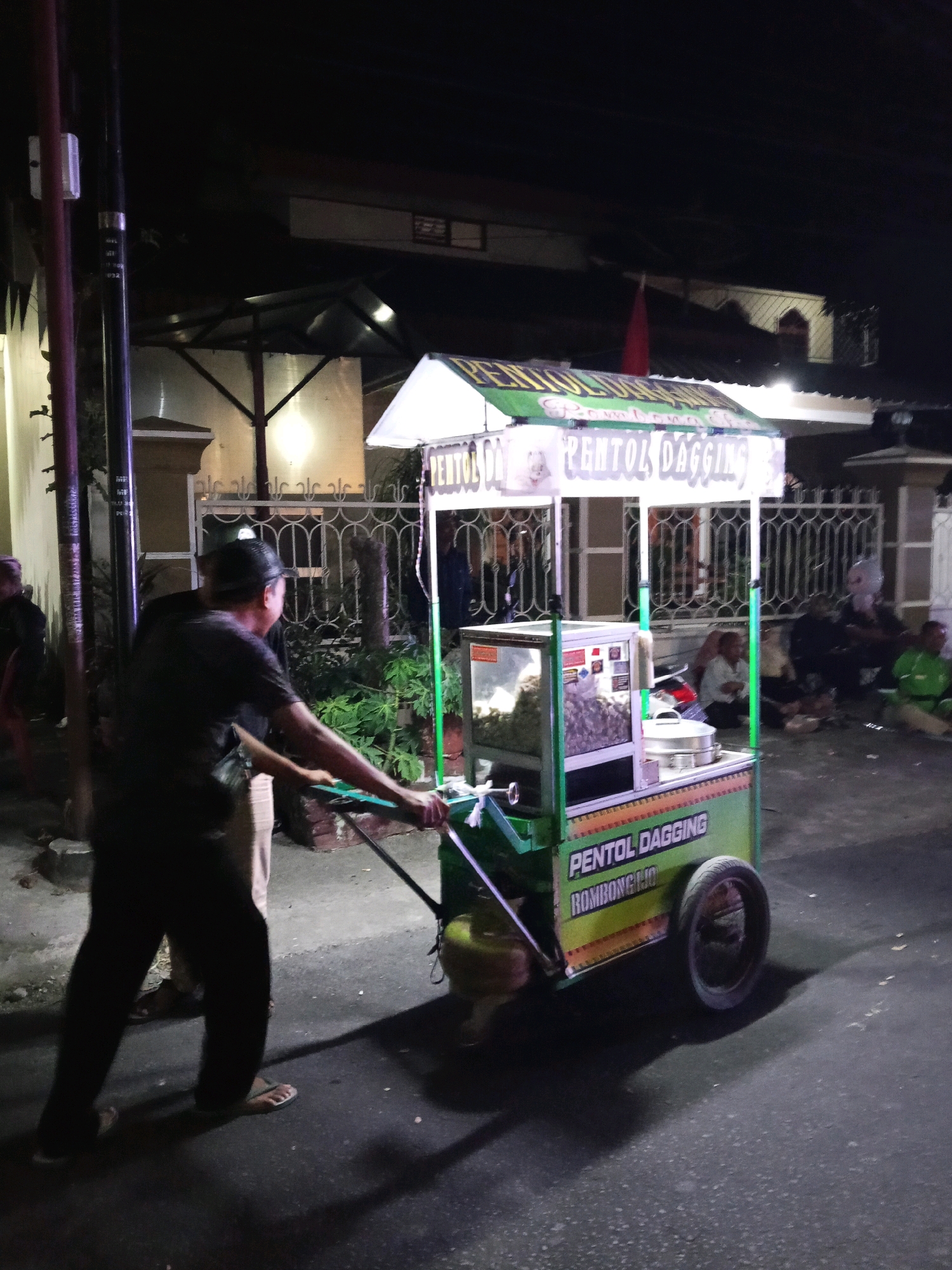 A man is pushing a food cart in the city of Bendosewu, Indonesia at night. The cart is green and white and has "PENTOL DAGGING" written on it in purple letters. There are people sitting and standing around the cart. The background shows a street with houses and a fence. The scene is illuminated by artificial lights.