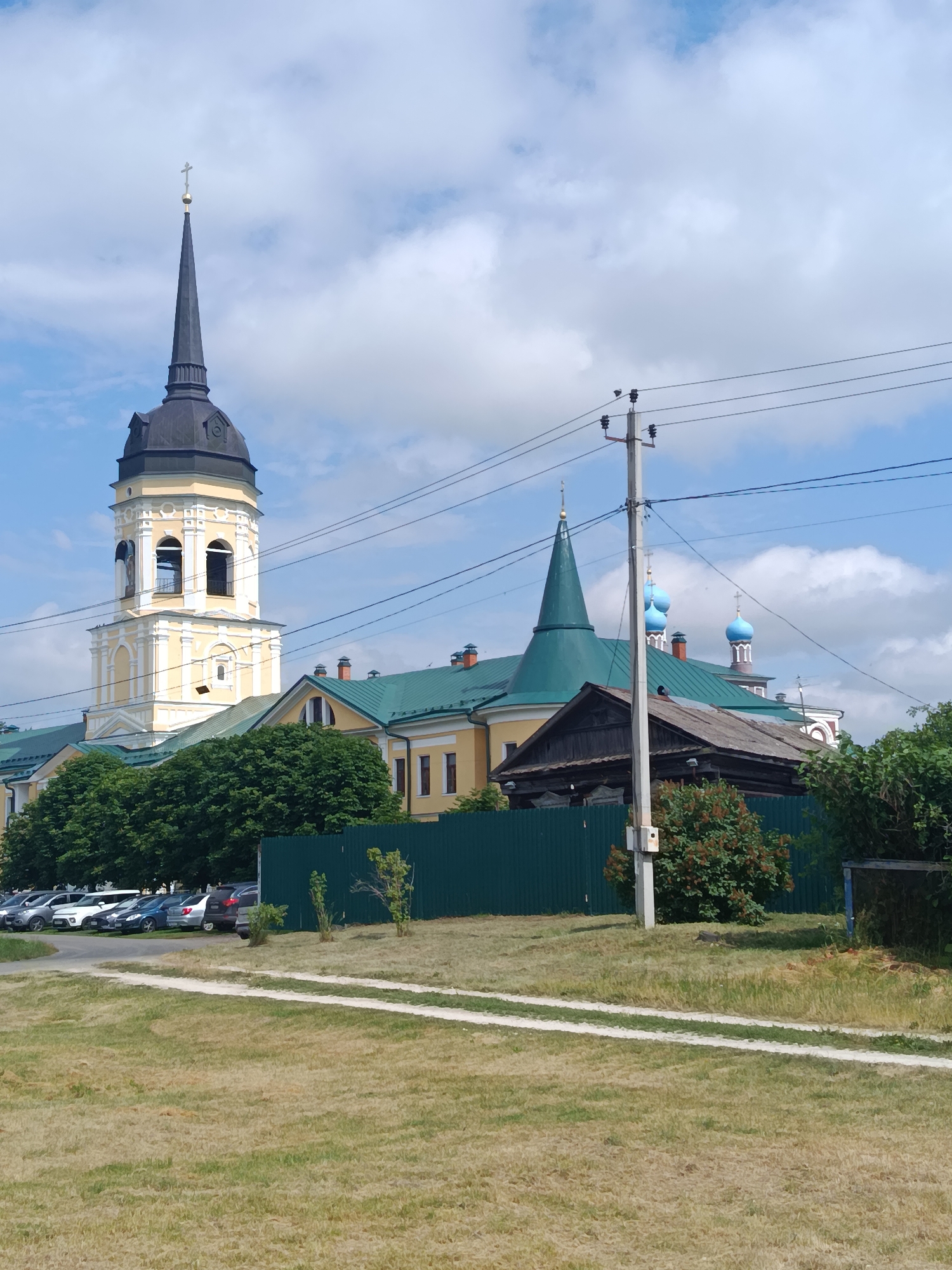The image depicts the Ascension Cathedral, a prominent religious site located in the city of Radovitsy, Russia. The cathedral complex features a tall, yellow bell tower with a dark spire topped by a cross, and several other buildings with green roofs. In the foreground, a gravel path is visible, with dry grass on either side. A dark green metal fence separates the cathedral grounds from the foreground area. Several parked cars are visible behind the fence, and a cluster of lush green trees lines the edge of the property. Power lines and a utility pole are present in the middle ground, with a small, weathered wooden structure visible behind the fence, to the left of a bush with red berries. The sky is blue with scattered white clouds.