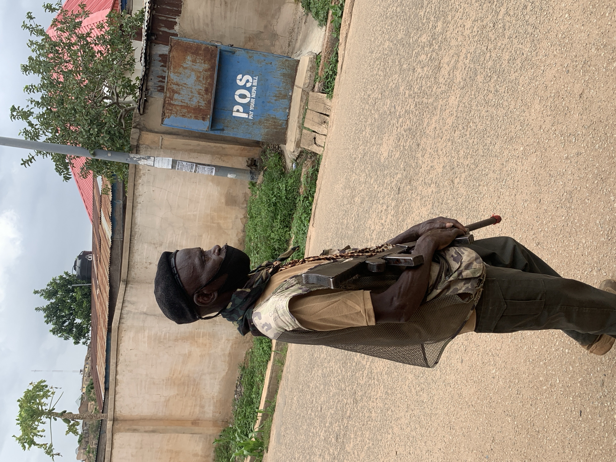 A man stands on a paved street, holding an AK-47 rifle. He wears a black beret, a black face mask, camouflage-patterned clothing, a khaki-colored shirt, dark green pants, and brown shoes. A mesh vest covers his chest. The rifle is held in both hands across his chest. Behind him is a wall, a blue metal box with the white letters "POS PAY YOUR NEPA BILL" on it, a tree, and a building with a red roof. The location is presumed to be Jos, Nigeria.