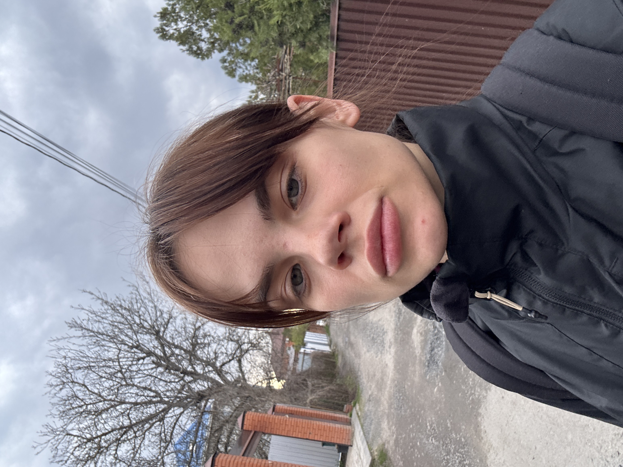 A young woman stands outdoors on what appears to be a city street. She faces the camera with a slight upward tilt. The background includes a gray sky, trees with bare branches, a brown corrugated metal fence, and brick buildings. The woman has brown hair and wears a black jacket and backpack. The setting is possibly located in Rostov-on-Don, Russia.