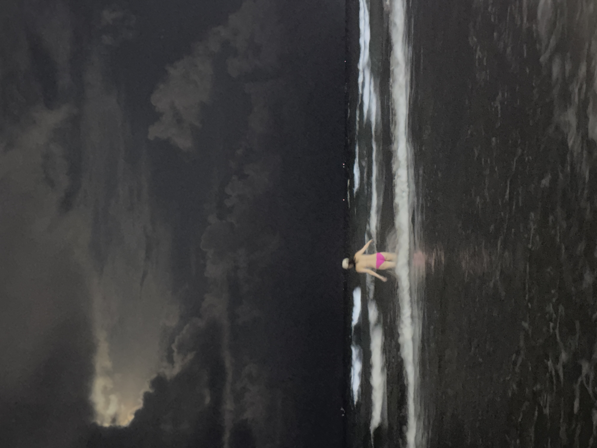 A lone individual wearing a pink bikini and a light-colored cap stands in the ocean at night. The person is facing away from the camera, with their arms extended to the sides. Gentle waves are breaking around them, and the water appears dark. The sky above is filled with dark, dramatic clouds, with a sliver of moonlight or distant light visible through a gap in the clouds on the left. In the distance, there are a few faint lights on the horizon, possibly from boats or buildings. The scene suggests a moody and atmospheric setting, possibly on a beach in Đà Nẵng, Vietnam, given the ocean and the potential for dramatic weather conditions.