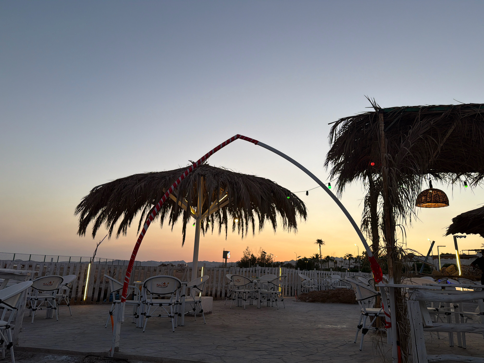 The image depicts an outdoor seating area of a beachside establishment at dusk, likely in Sharm El Sheikh, Egypt. The foreground features several white metal tables and chairs, some with the brand name "SADIKI" visible. Palm frond umbrellas provide shade, with one notable umbrella supported by a red and white striped pole. String lights with green bulbs are visible above, and a woven pendant light hangs from one of the palm fronds. The ground is paved with textured stone tiles.
In the background, a white picket fence lines the perimeter of the seating area. Beyond the fence, the landscape is sparsely populated with some structures, palm trees, and distant hills. The sky exhibits a gradient of colors, transitioning from a pale blue at the top to a warm orange and yellow near the horizon, indicating the setting sun. The overall atmosphere is serene and hints at a place of relaxation and dining.