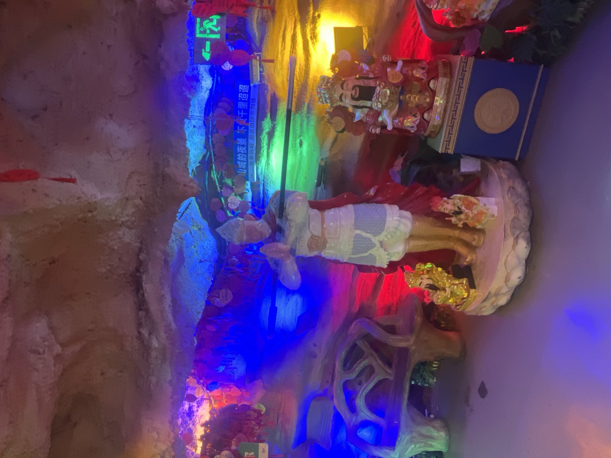 The image captures an indoor, cave-like environment bathed in a vibrant array of artificial lights, predominantly blue, red, green, and yellow. In the foreground, a large statue, possibly depicting a deity or warrior, stands adorned in white and gold, holding a staff. Behind it, a smaller, ornate statue of a bearded figure with elaborate headwear and a red robe is visible, positioned next to a blue box featuring a gold circular emblem.
A glowing green exit sign, showing a person running, is prominently displayed on the left wall. Below it, a banner with Chinese text is visible. The OCR text translates to: "山城的夜景不慢干里迢迢" (The night view of the mountain city is not slow for thousands of miles) and "出来玩呀反正也挣不到钱" (Come out and play, anyway, you won't earn money), suggesting a leisure or tourist attraction.
The setting features rough, textured walls resembling natural rock formations, enhancing the grotto or cave atmosphere. Small red decorative elements, possibly lanterns or artificial foliage, are interspersed throughout the scene. The dynamic and colorful lighting creates a mystical and engaging ambiance for this cultural display, likely located in Yuzhong District, China. The scene suggests a well-illuminated exhibition designed to captivate visitors.