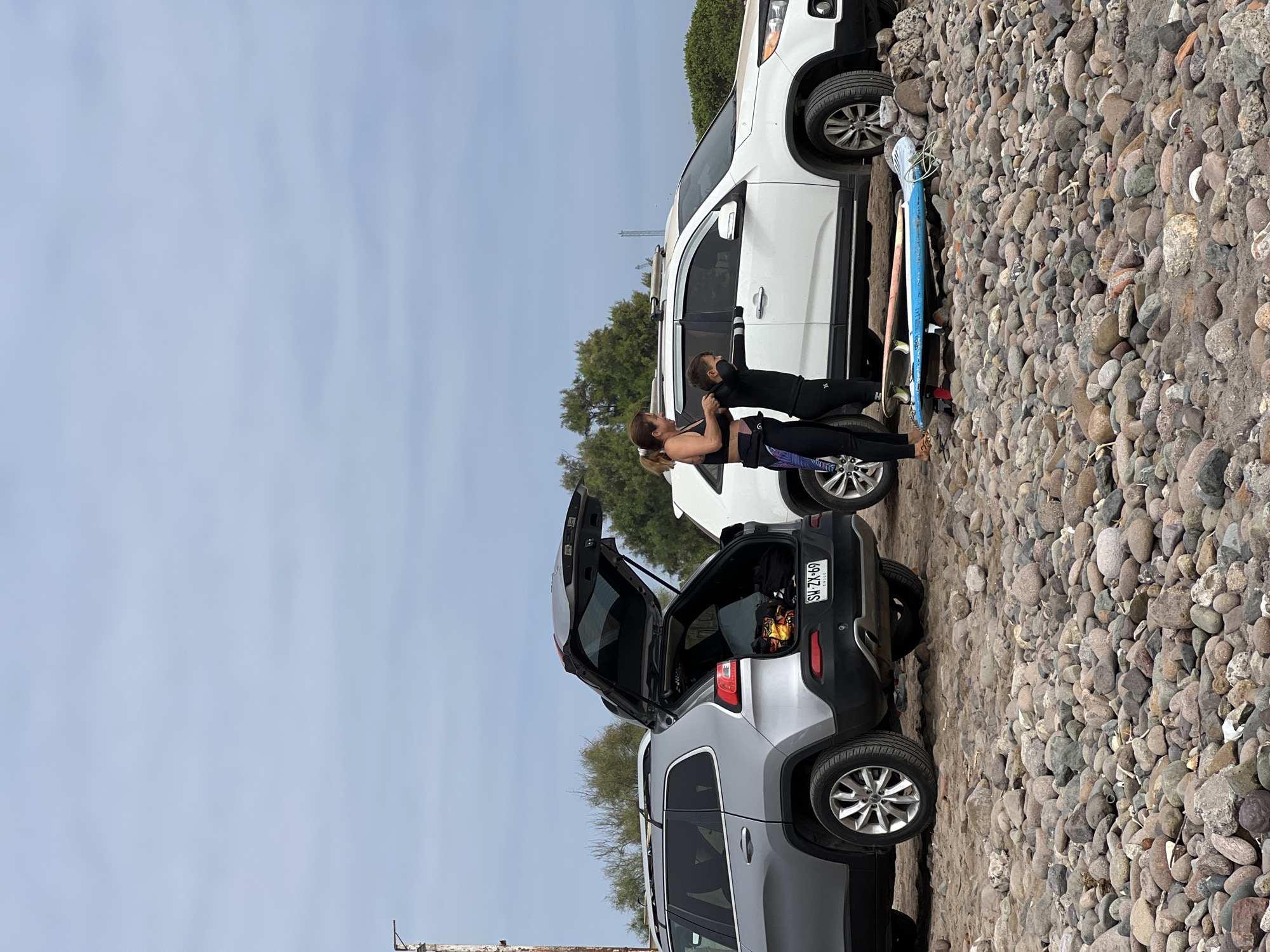 The image depicts a pebble-strewn area, possibly a beach or a parking area near a beach. Two vehicles are parked: a light gray SUV with its trunk open and a white SUV. A light blue surfboard leans against the white SUV. A woman and child dressed in what appears to be athletic or swimwear stand between the vehicles. The woman appears to be attending to the child. The license plate on the gray SUV is visible, reading "SW ZL 69". The location is likely Arica, Chile, given the instruction.