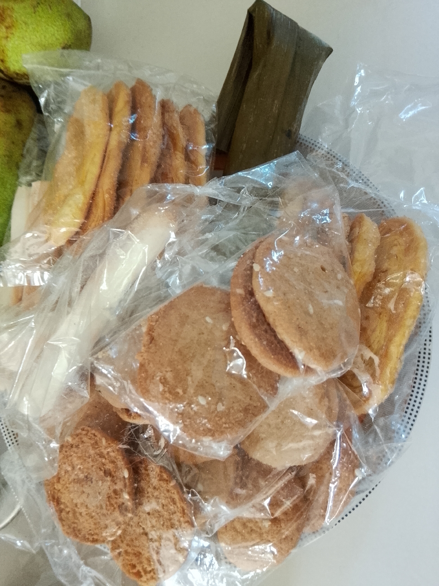 The image displays a collection of various packaged Filipino snacks, likely from Davao City, Philippines, resting in a shallow container with a black and white checkered rim.
The primary subjects are several types of food items, each individually sealed in clear plastic bags. In the upper left, out of focus, are two large, green-skinned fruits, possibly avocados or unripe mangoes.
Within the container, there are at least three distinct types of snacks:
1. **Golden Brown Strips:** Several bags contain long, finger-like, golden-brown snacks, which appear crispy or flaky, possibly similar to *lubid-lubid* or *shing-a-ling*.
2. **Round Flat Cookies/Crackers:** Multiple bags hold light brown, round to oval-shaped flat snacks, resembling cookies or crackers, such as *otap*. Some of these appear to have specks of sugar or other toppings on their surface.
3. **Leaf-Wrapped Item:** Towards the top right, a single item is distinctly wrapped in a dark green, folded banana leaf, typical for traditional Filipino rice cakes like *suman*.
The snacks are arranged somewhat haphazardly within the container, suggesting they are either freshly bought, prepared for serving, or awaiting storage. The clear plastic packaging highlights their homemade or artisanal appearance. The background consists of a plain, light-colored surface, likely a table or countertop. No people are visible, and there is no discernible text on any of the packaging. The setting appears to be indoors, possibly a home kitchen or a small food stall.