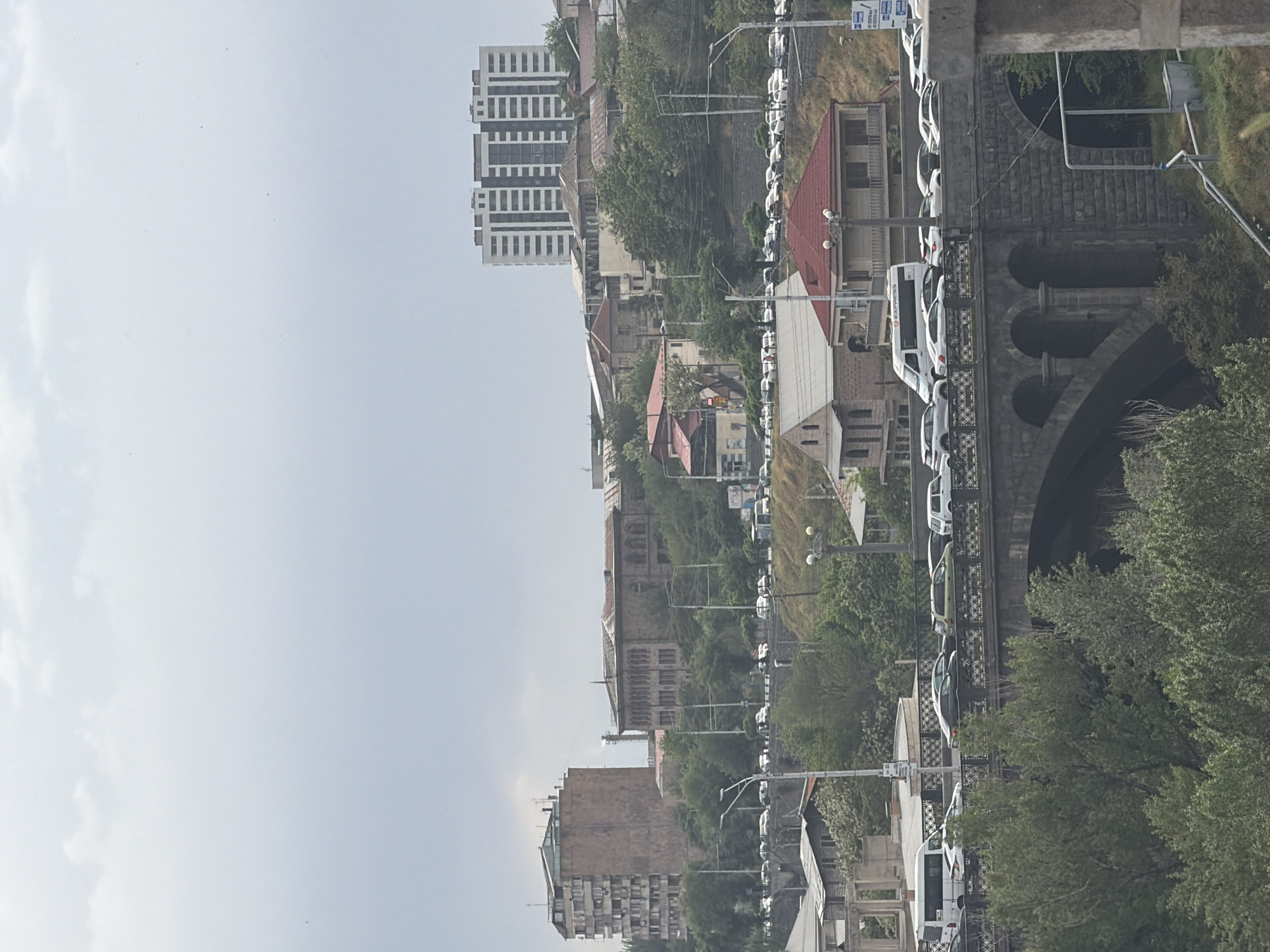 This is an outdoor shot of Yerevan, Armenia. The scene features a stone bridge, likely a viaduct, with vehicles parked or moving across it. The architecture includes stone buildings with red-tiled roofs, and modern high-rise apartment buildings in the background. Lush green trees and foliage surround the structures, suggesting a combination of urban and natural landscapes. The sky is overcast, with a soft, diffused light. The image appears to be taken from an elevated viewpoint, possibly from another building or elevated terrain.